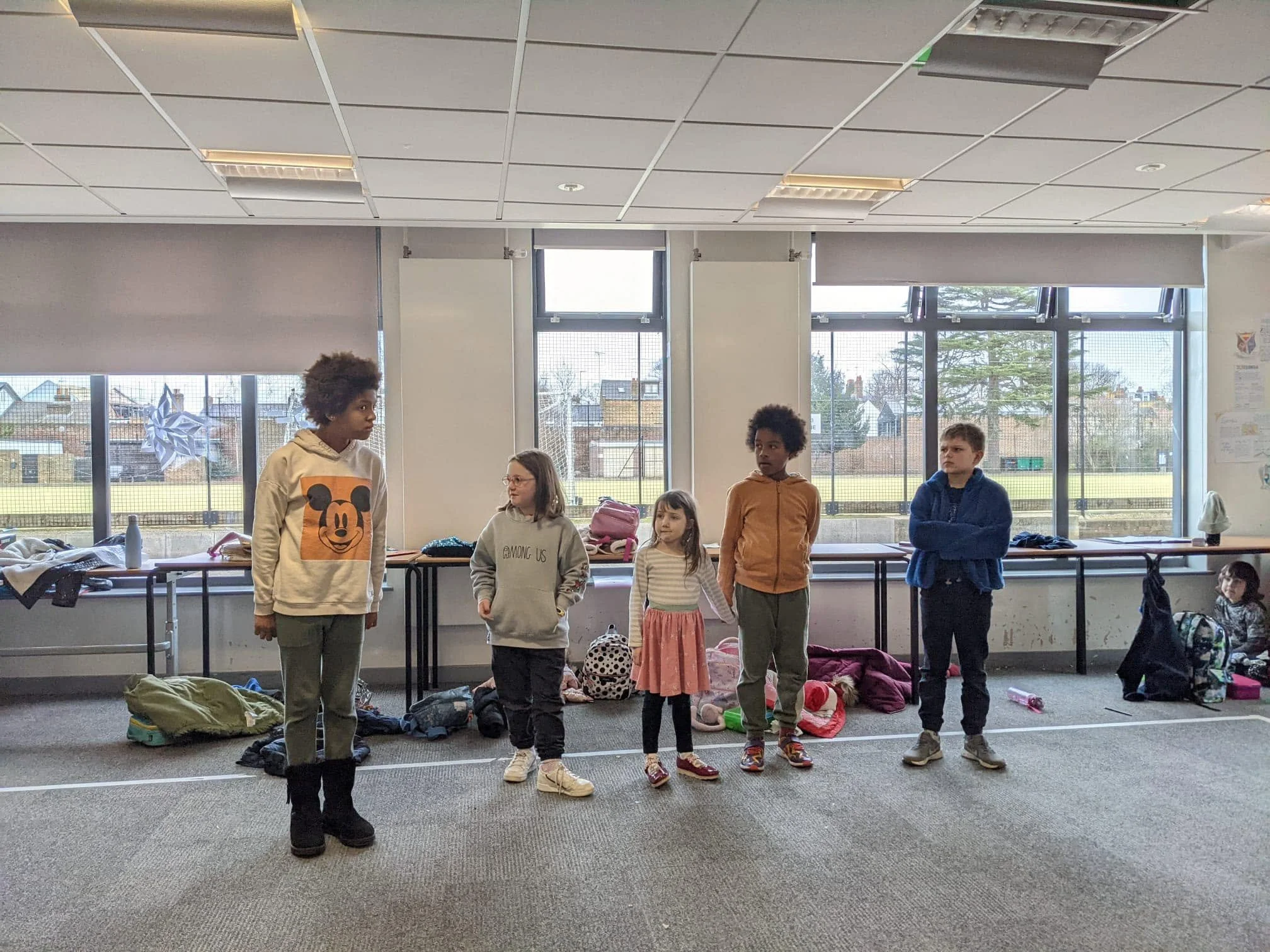 Children and teenagers standing in a room with large windows, surrounded by scattered backpacks and belongings, likely in a school or community center.