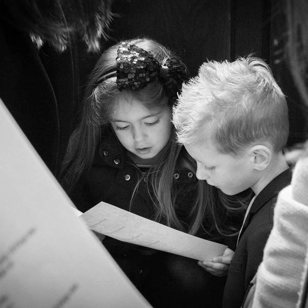 Two children, a girl with a bow in her hair and a boy, are looking at a piece of paper together. They are in a group setting with other people partially visible around them.