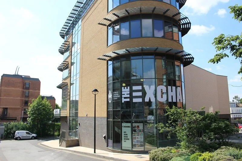 Modern building with large glass windows and the word 'THE EXCHANGE' displayed on the front, surrounded by trees and a parking lot.