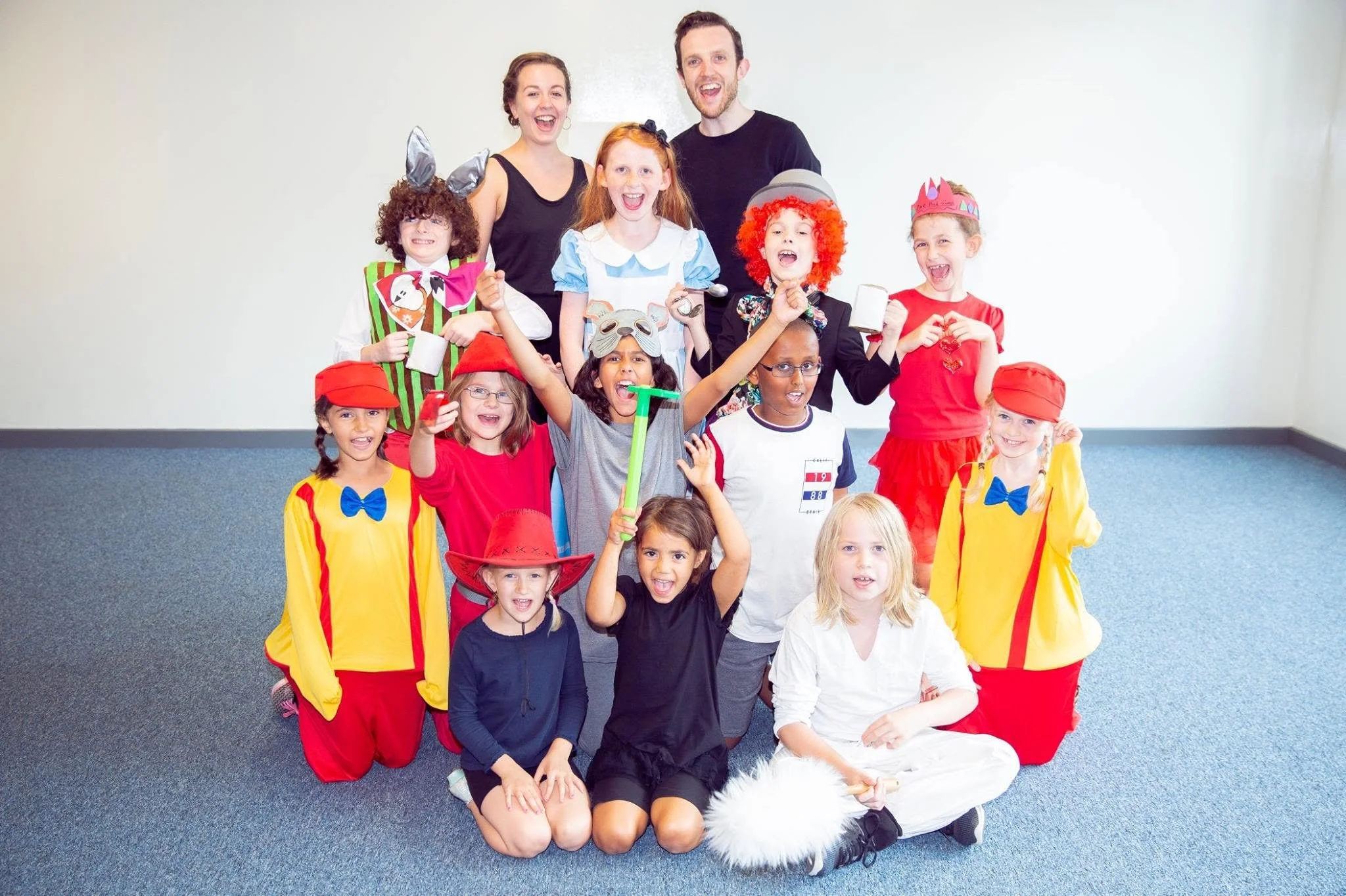 Group of children and adults in colorful costumes celebrating together in a room with plain walls and carpeted floor.