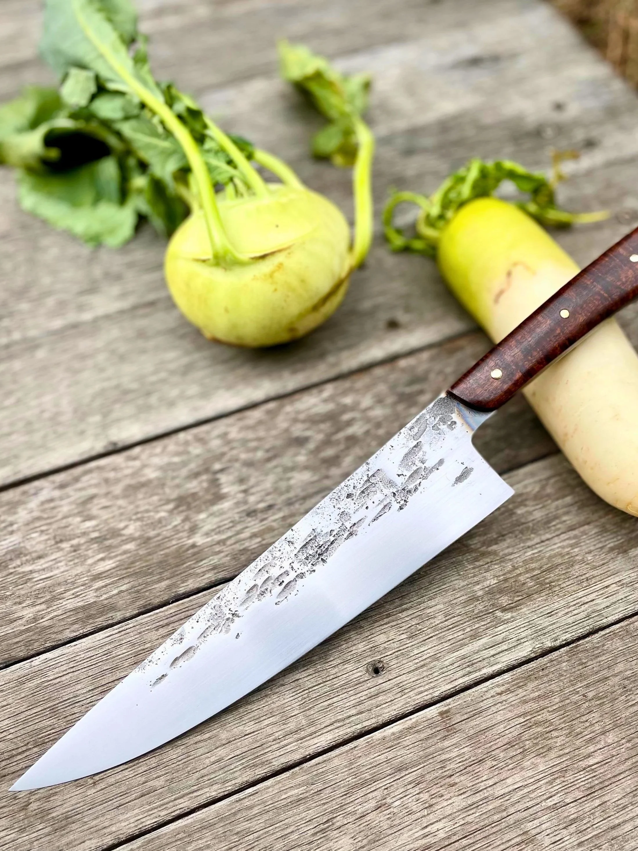 Two radishes with green leaves on a weathered wooden surface, and a stainless steel knife with a brown handle nearby.
