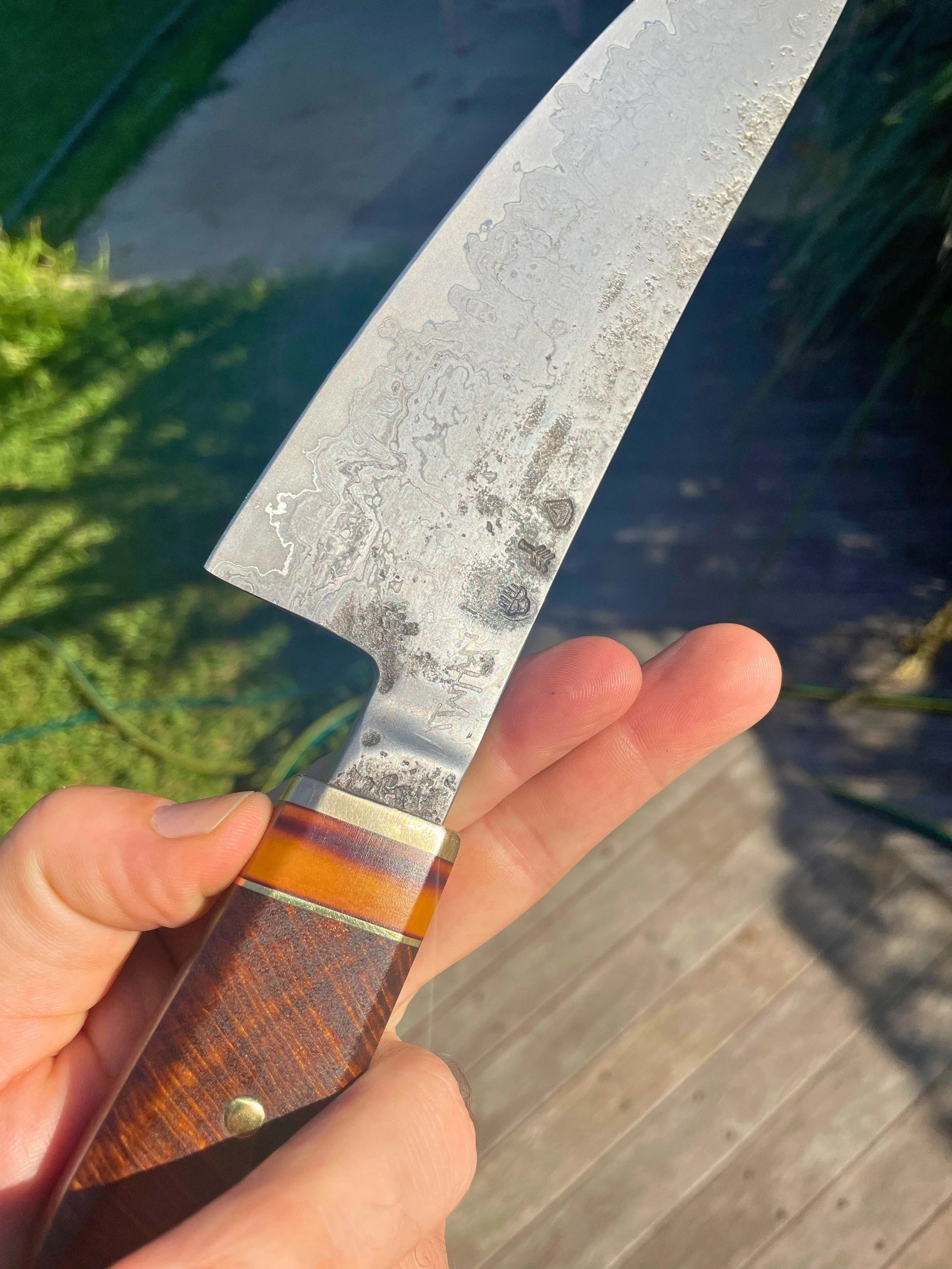 Close-up of a person's hand holding a damascus steel chef's knife with a wooden handle over a wooden surface, with greenery in the background.