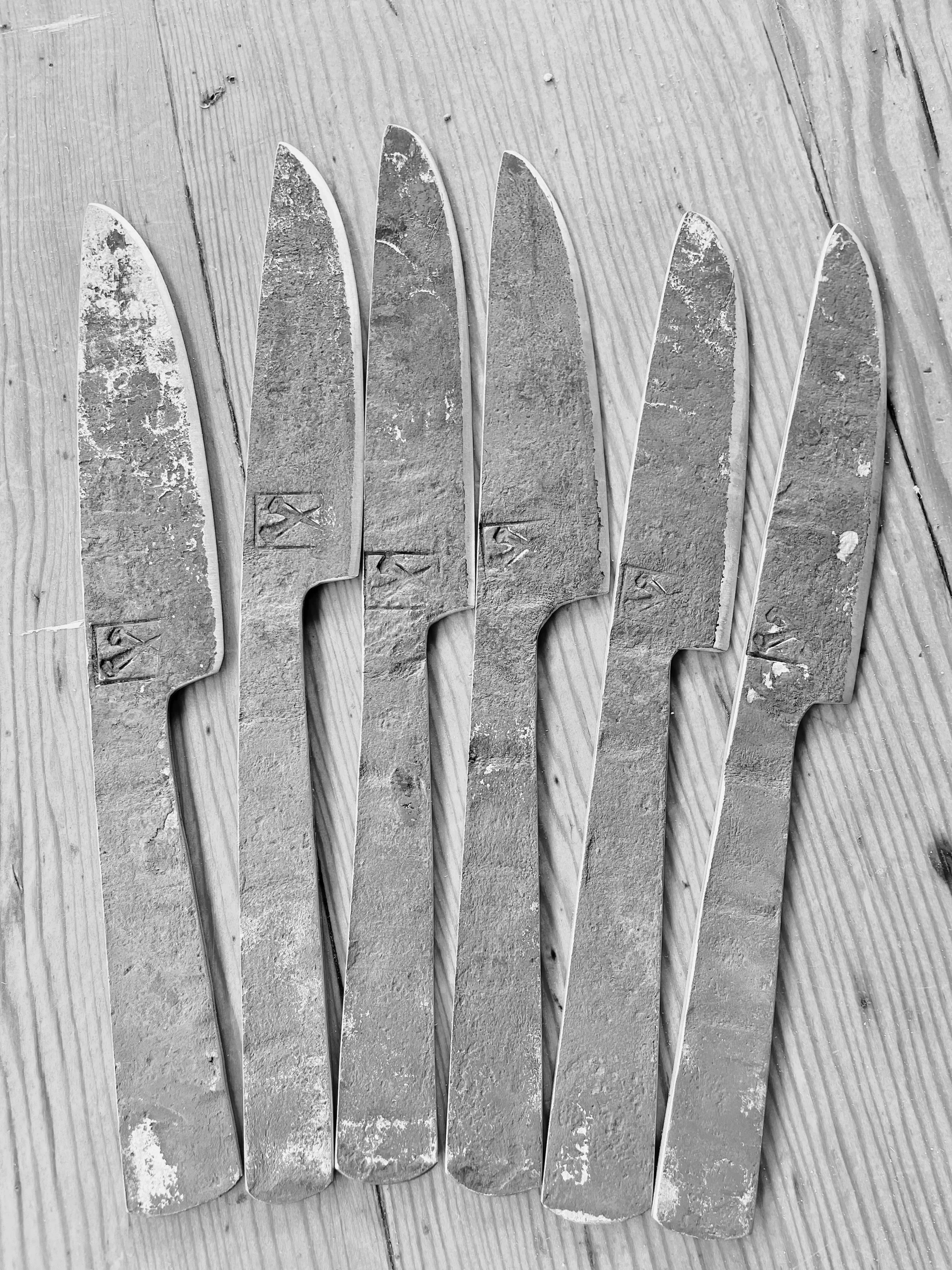 Six rustic hand forged butter knives, arranged side by side on a wooden surface.