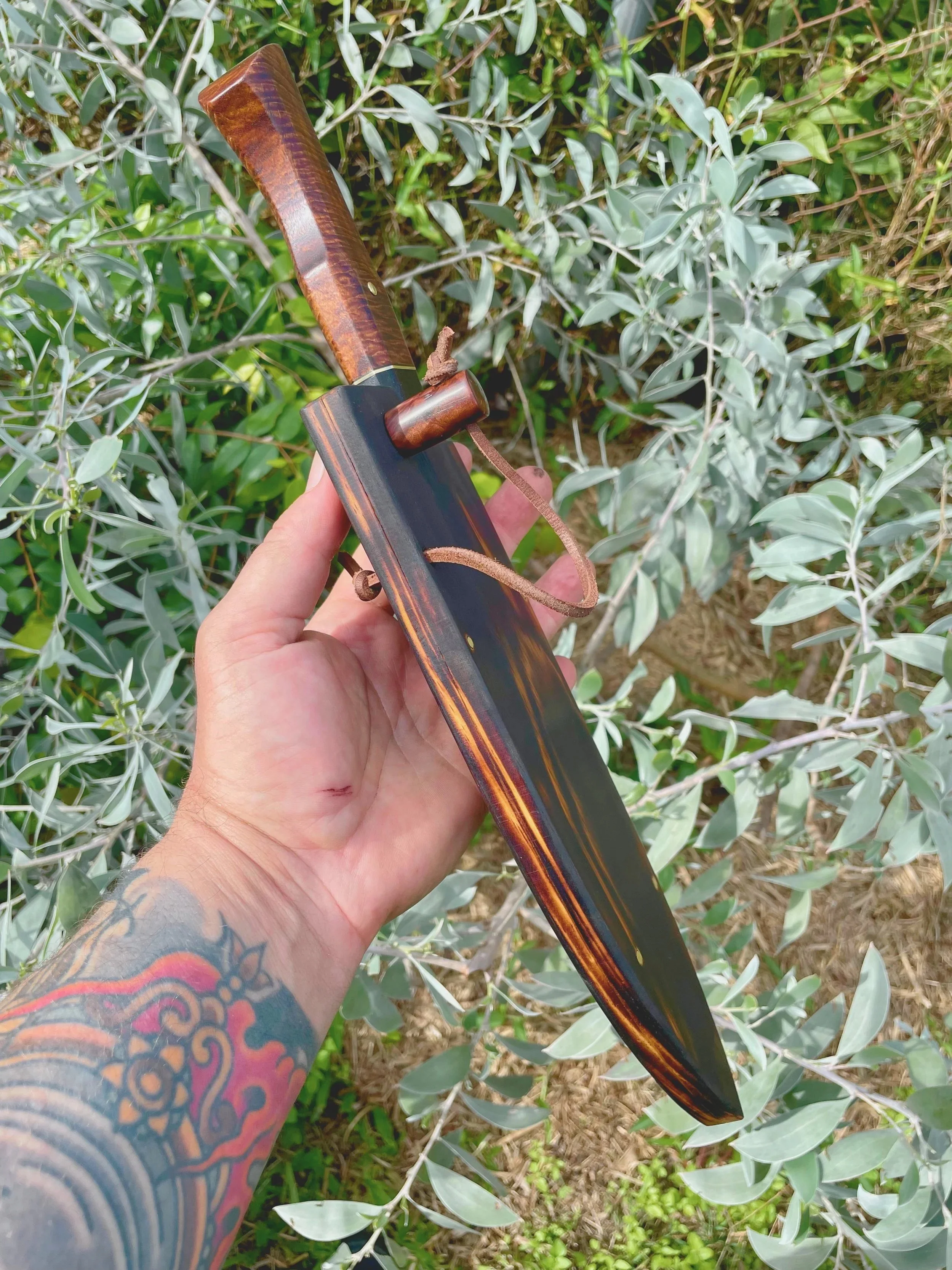 A tattooed arm holding a knife with a wooden handle, seen against a background of green and silver leaves.