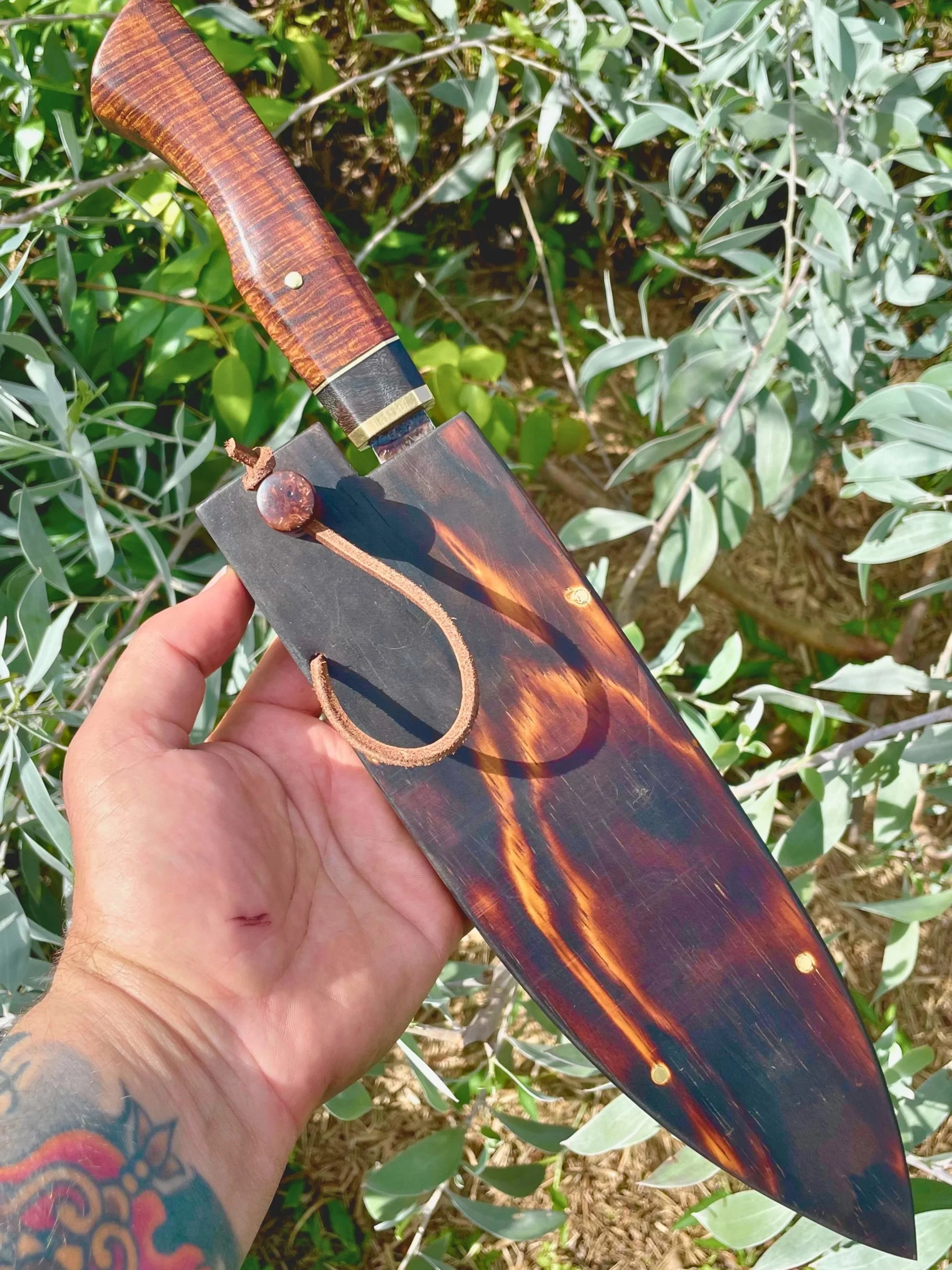 Close-up of a bow held in a person's hand outdoors among green foliage, holding a chef knife in a hand carved wooden sheath.