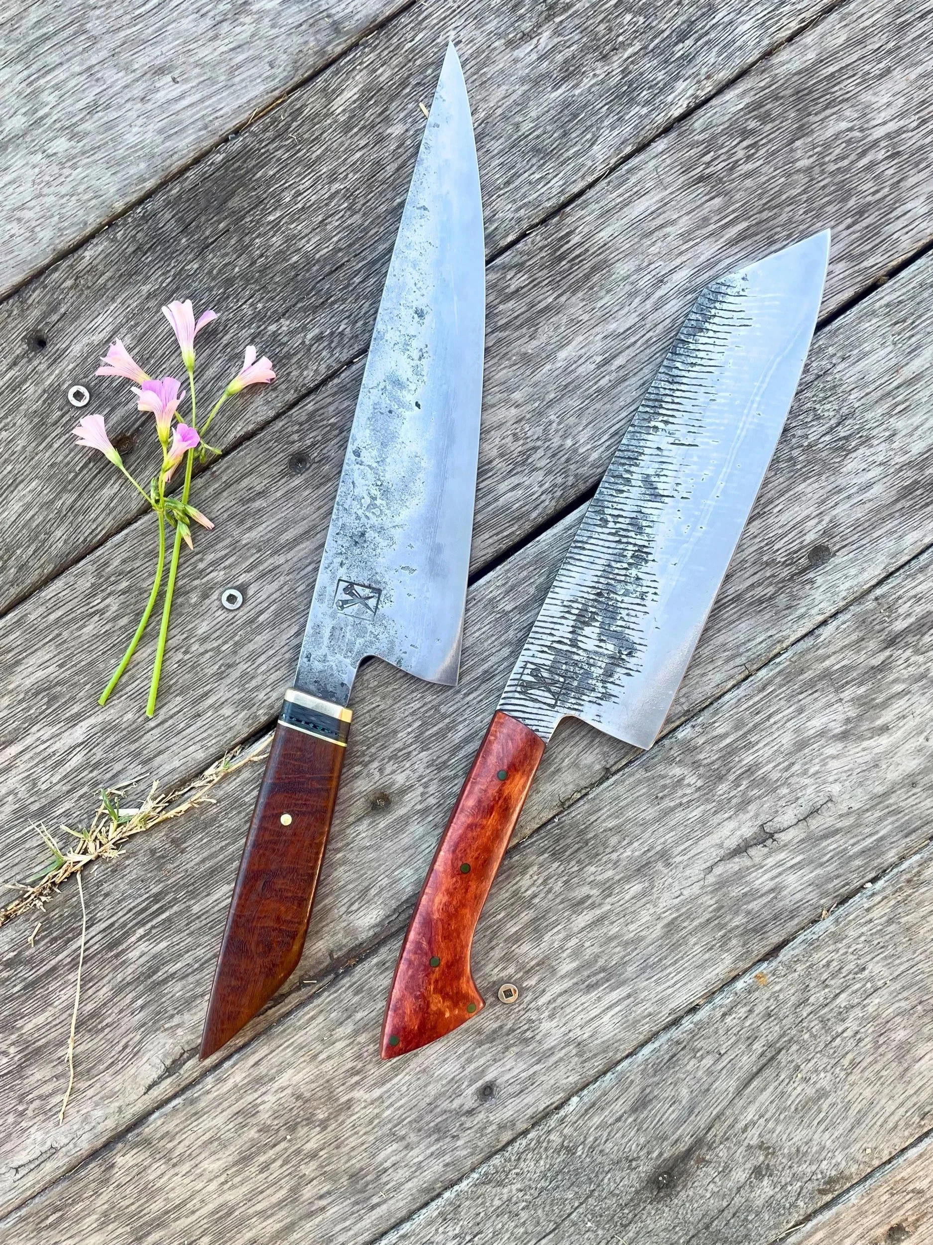 Two large kitchen knives with wooden handles and a bunch of pink flowers on a weathered wooden surface.
