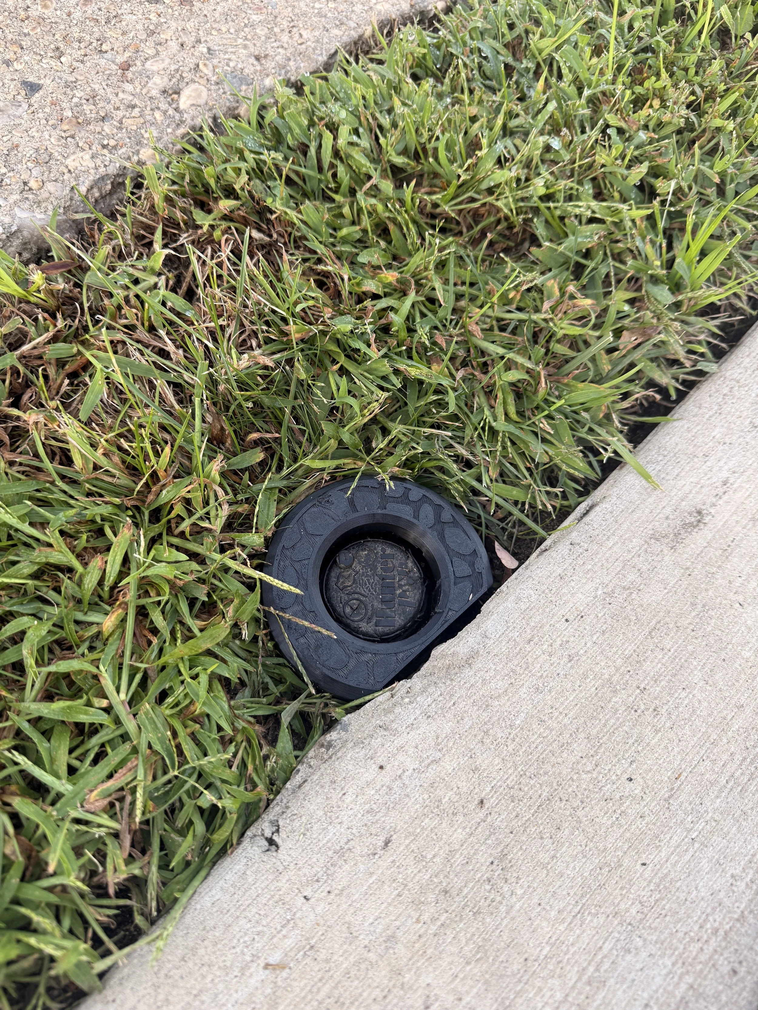 Black IngenioTech Edge Guard installed around a sprinkler rotor where lawn grass meets a concrete path.