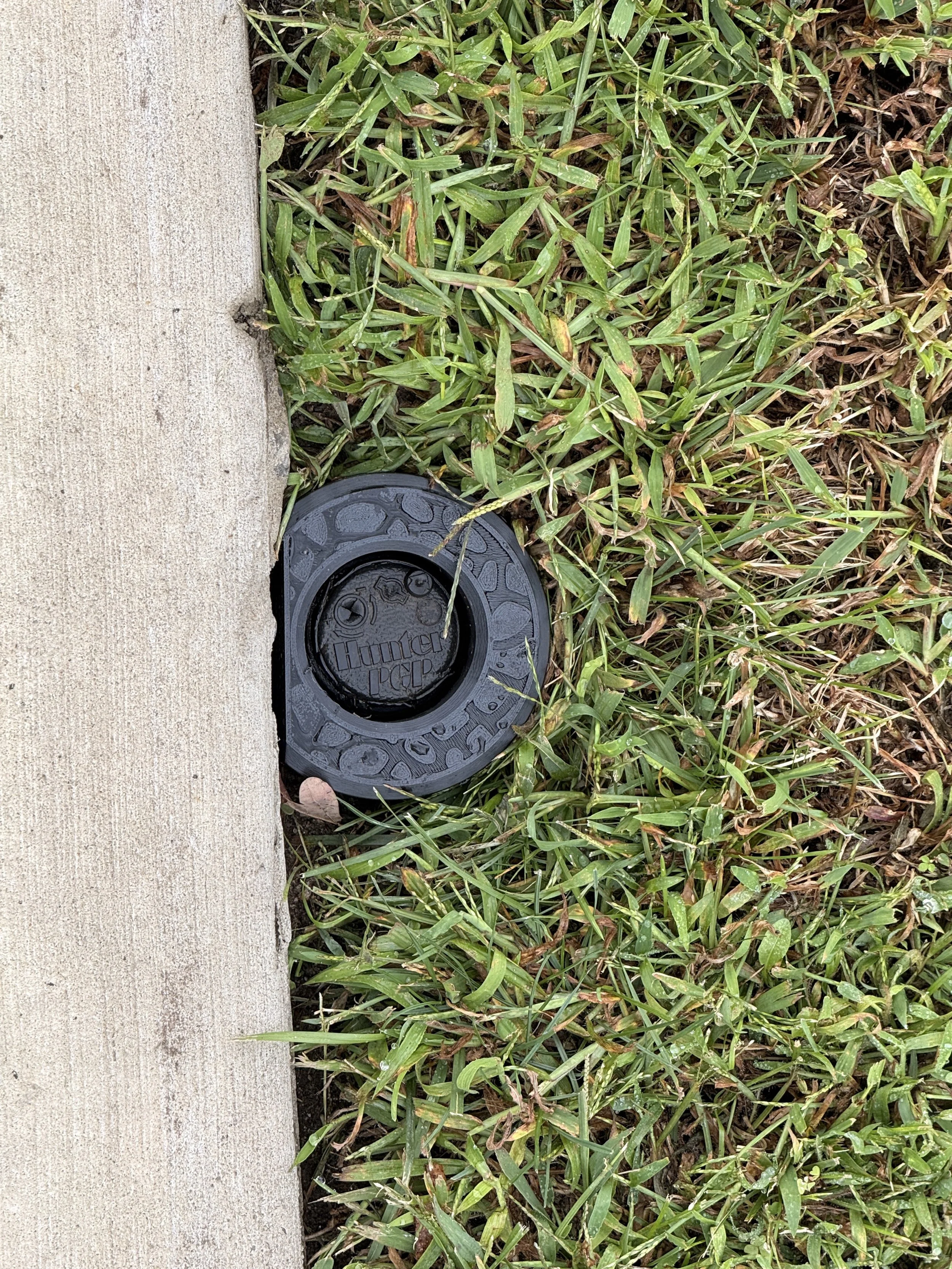 Top view of a black IngenioTech Edge Guard around a Hunter PGP rotor, with its flat side beside a straight concrete border.