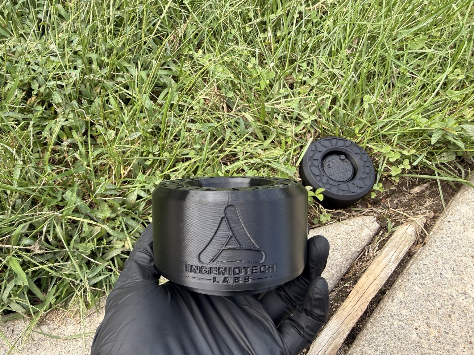 A black IngenioTech Labs sprinkler guard held beside a lawn installation