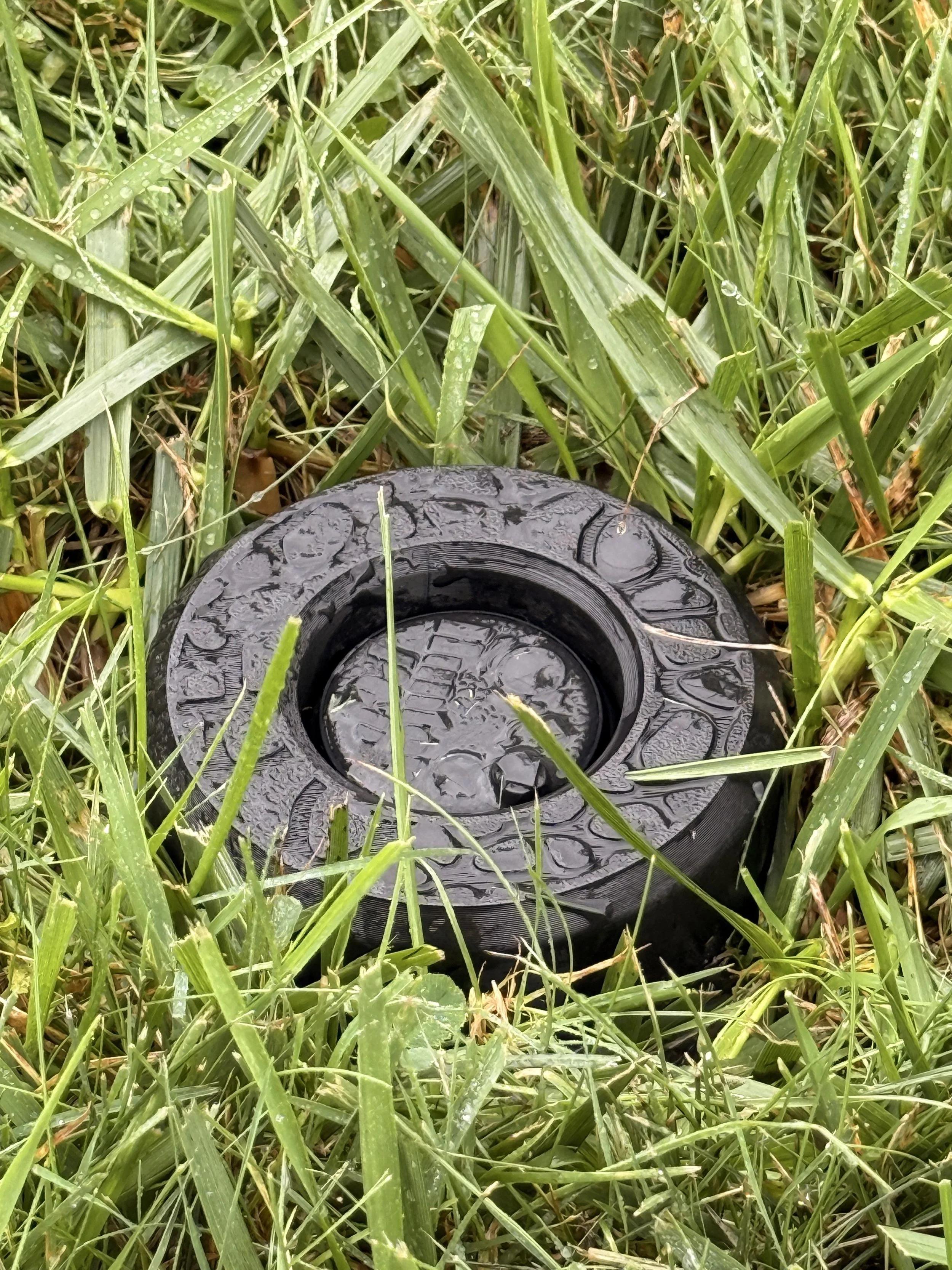 Close-up of a black IngenioTech Full Guard installed around a sprinkler rotor in wet lawn grass.