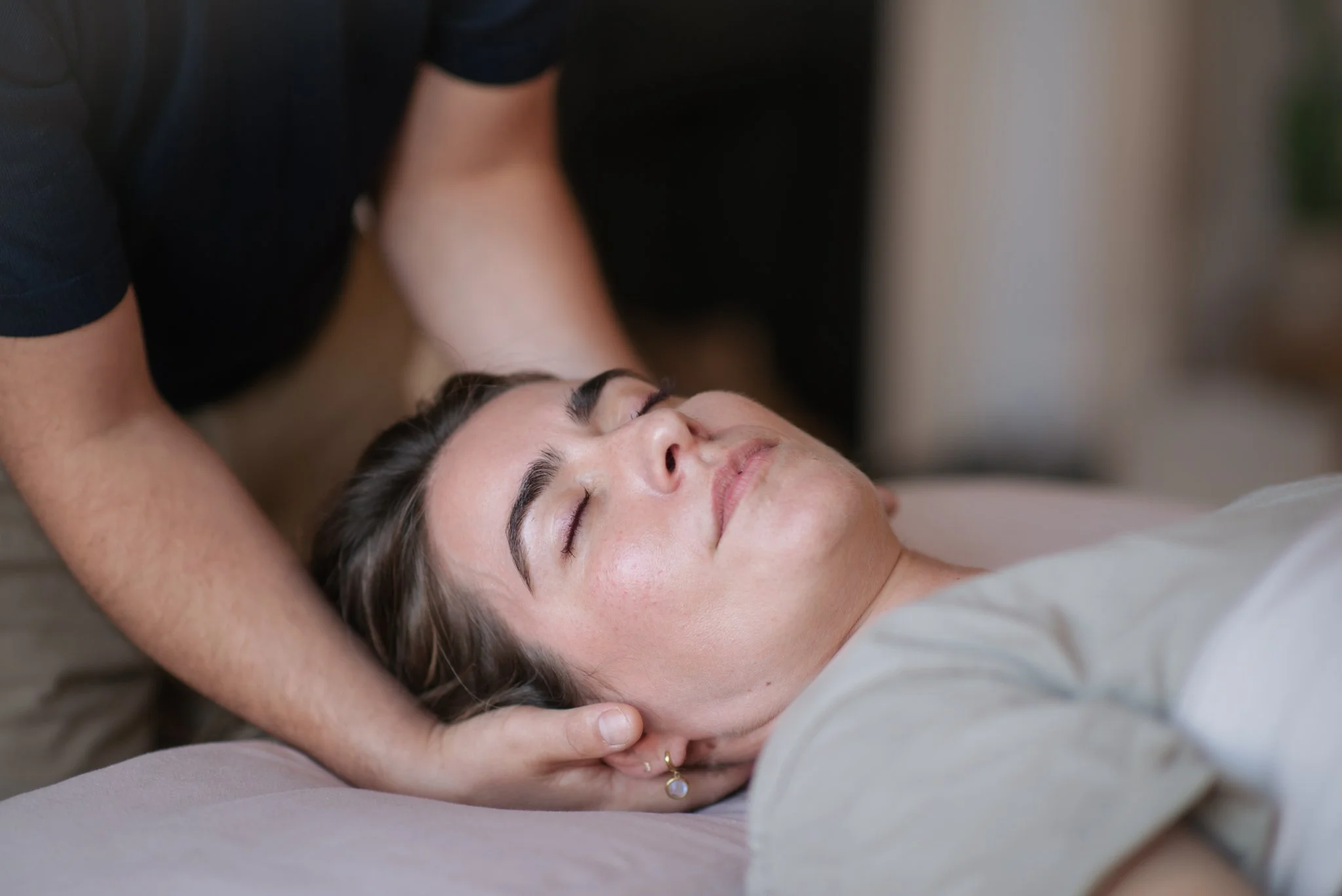 A woman lying on a massage table with her eyes closed, receiving a massage, with a massage therapist gently supporting her head.