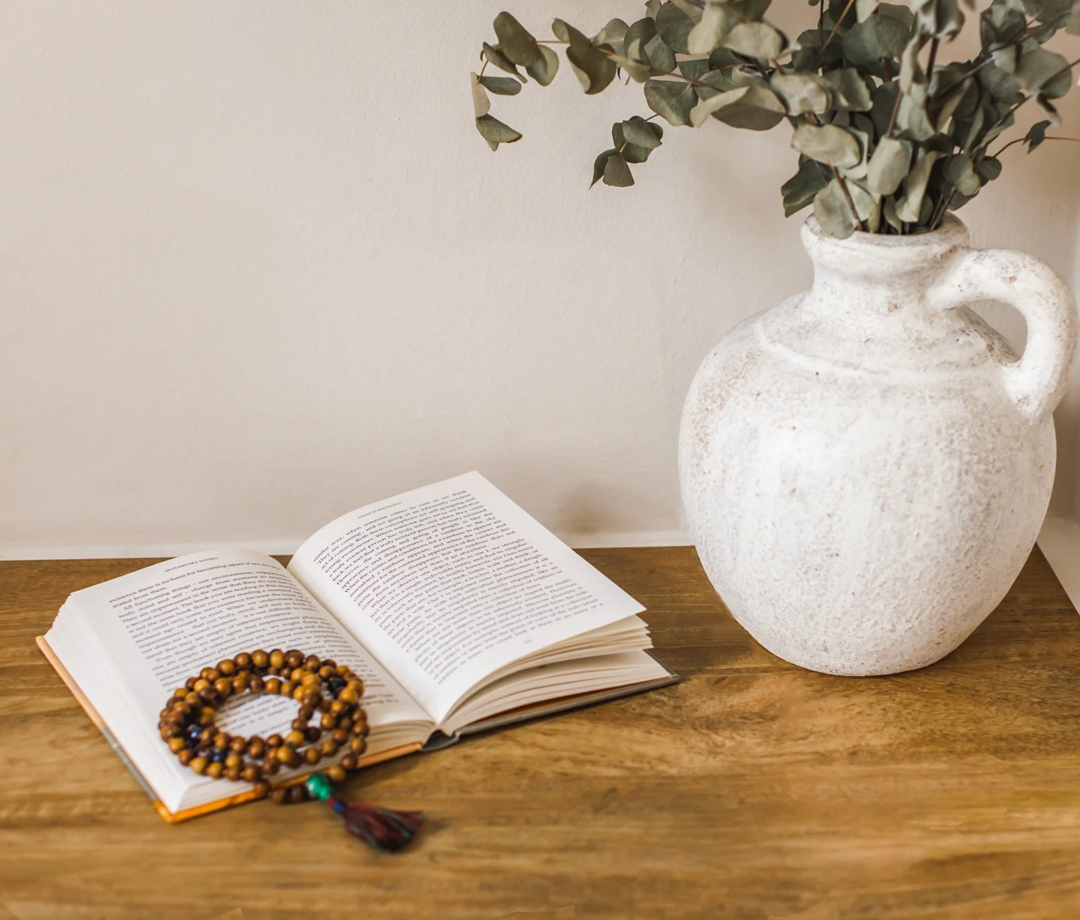 A wooden surface holding an open book, a wooden beaded bracelet, a white textured ceramic vase with a handle, and dried eucalyptus leaves.
