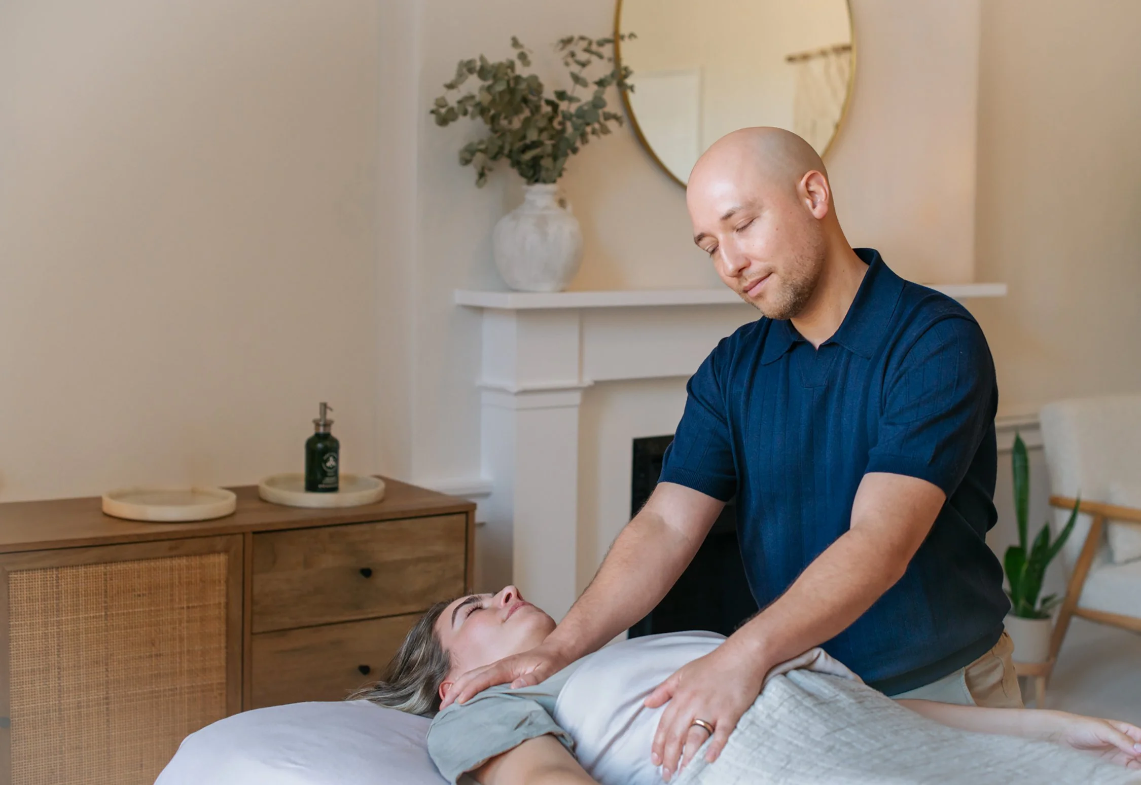 A woman receiving a massage from a male therapist in a cozy, well-decorated room with a round mirror, a ceramic vase with greenery, and a wooden cabinet.
