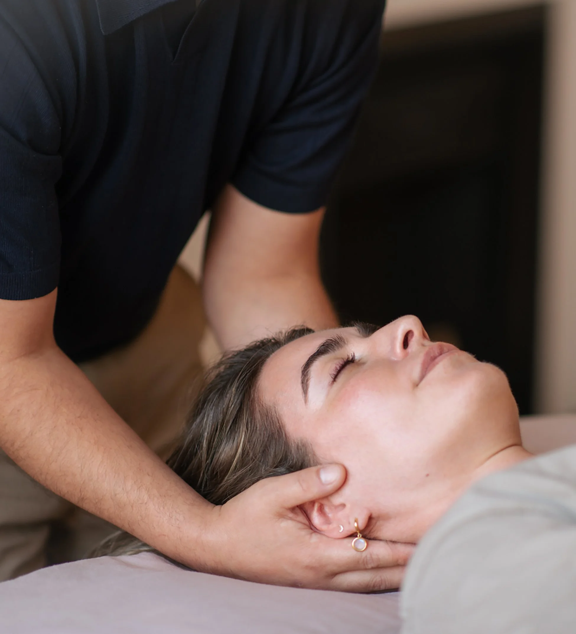 A woman with closed eyes receiving a massage or chiropractic adjustment, lying on her back, with a person's hand on her neck and shoulder area.