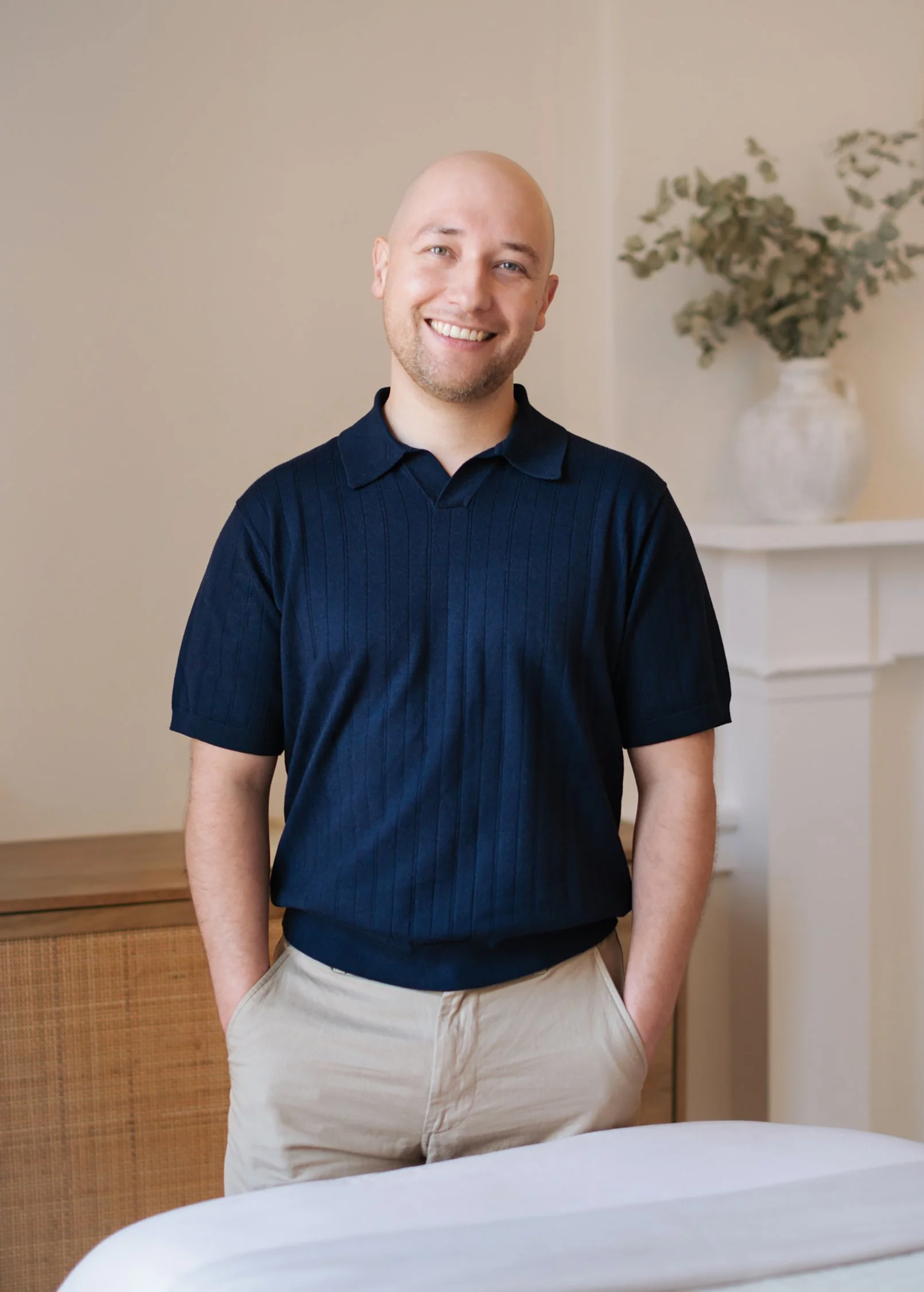 A smiling man with a bald head and light skin, wearing a dark blue shirt and beige pants, stands indoors in front of a beige wall and a white dresser with a vase of green leafy plants.