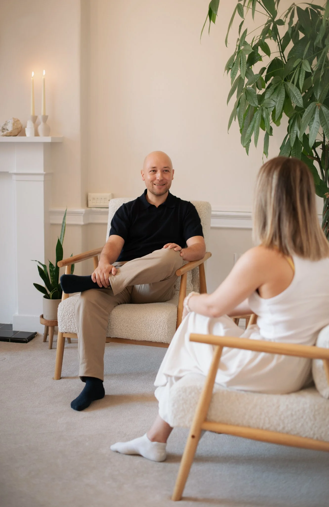 A man sitting on a chair engaging in conversation with a woman who is sitting on a similar chair. The setting is a cozy, well-lit room with a tall indoor plant, a fireplace mantel with two lit candles, and some decorative objects. Both individuals appear relaxed and smiling.