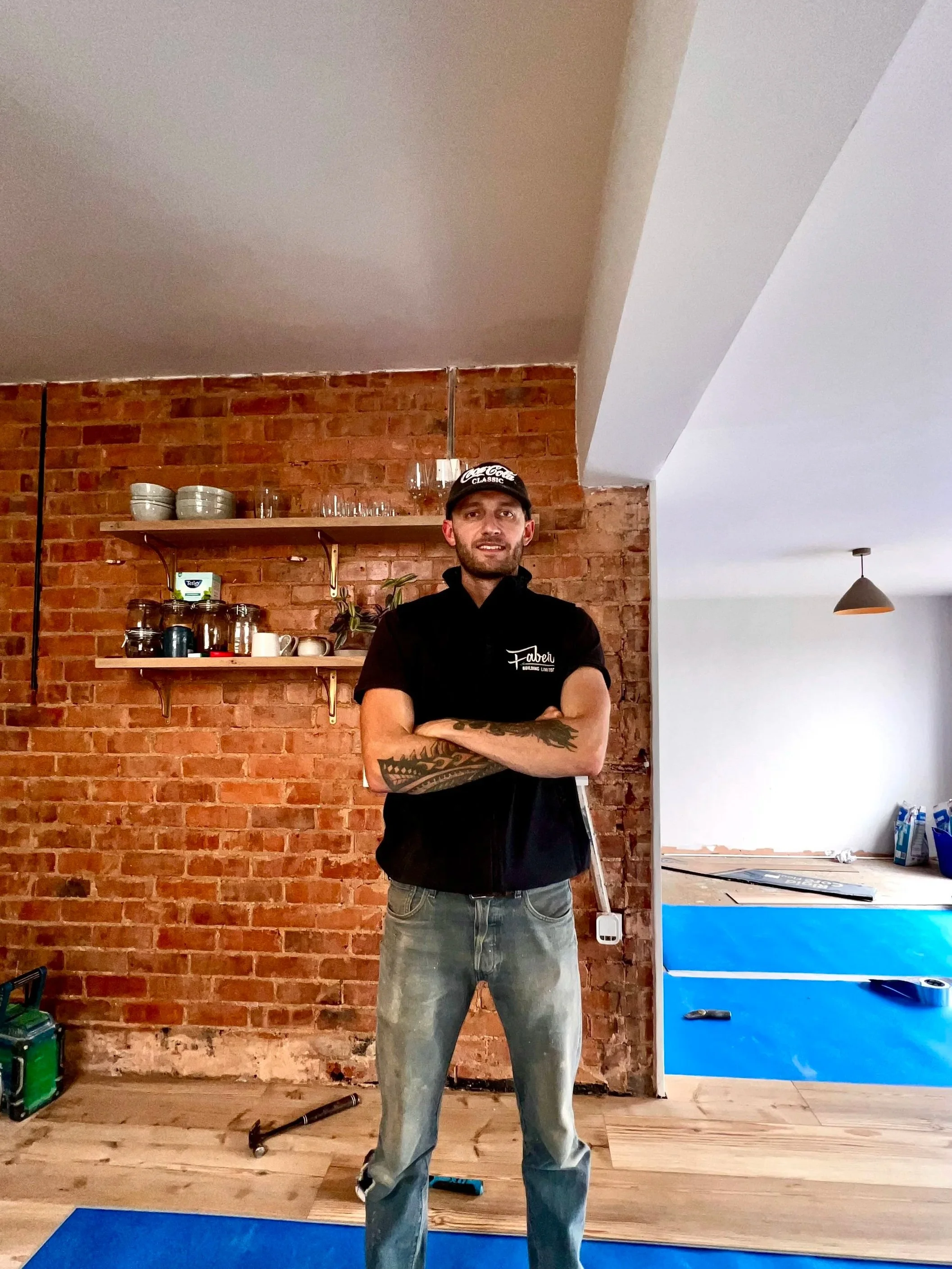 A man with tattoos on his arms wearing a black cap and black shirt, standing in a room with a brick wall, wooden flooring, and a blue exercise mat.