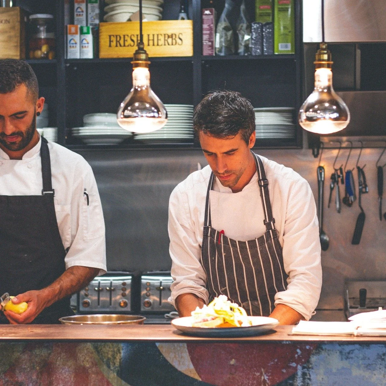 Two male chefs in striped aprons preparing food in a professional kitchen with hanging light fixtures, shelves with dishes, and utensils on the wall.