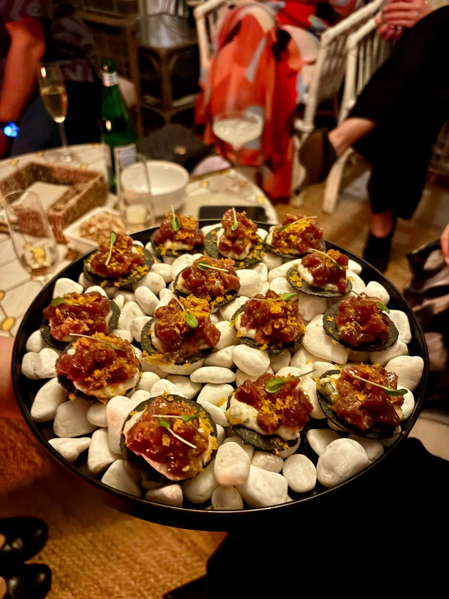Close-up of a tray of appetizers with small round crackers topped with a spicy meat mixture and garnished with small green leaves, resting on white stones. In the background, a table with various food items and drinks, including a glass of white wine and a green bottle, is visible, surrounded by people at a social gathering.