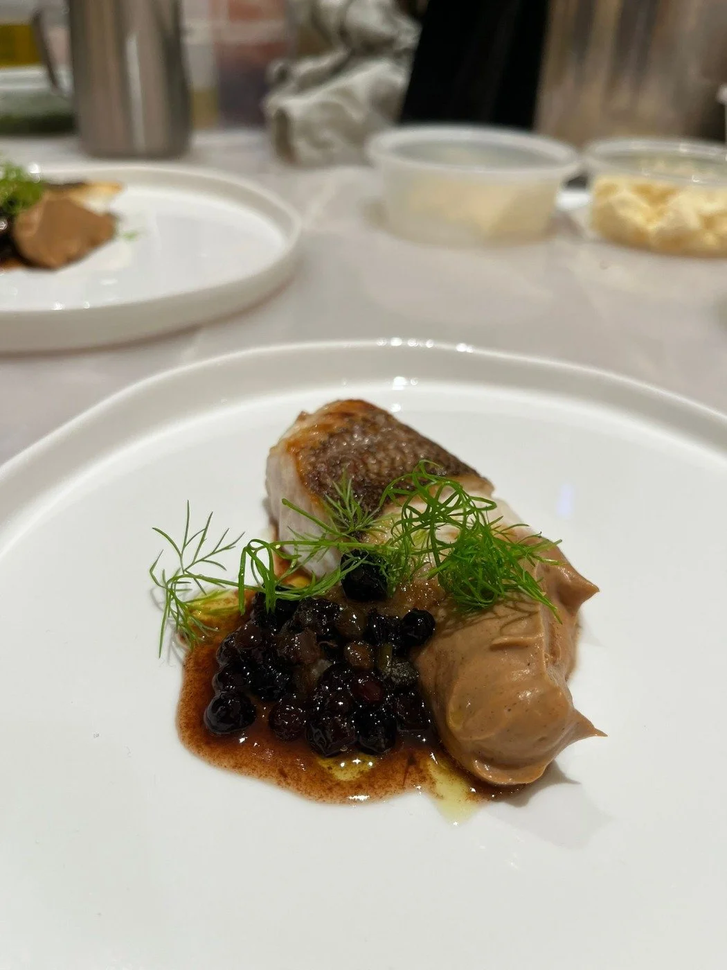 A plated portion of gourmet food with a piece of meat, a dollop of creamy sauce, garnished with greens and black berries on a white plate.