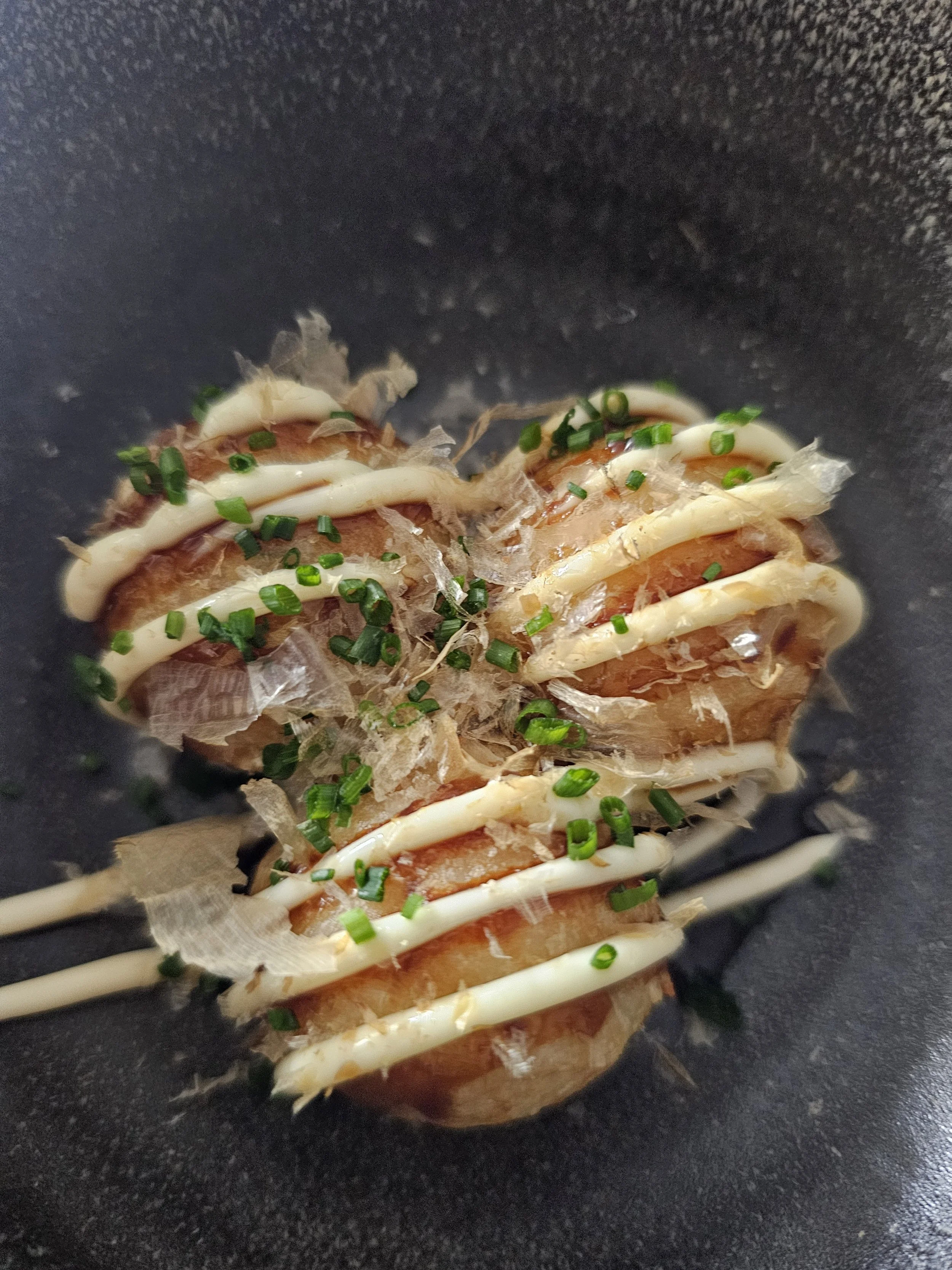 Close-up of a food dish with thinly sliced meat or fish, topped with chopped green onions, drizzled with mayonnaise and possibly soy sauce, arranged on parchment paper in a black bowl.