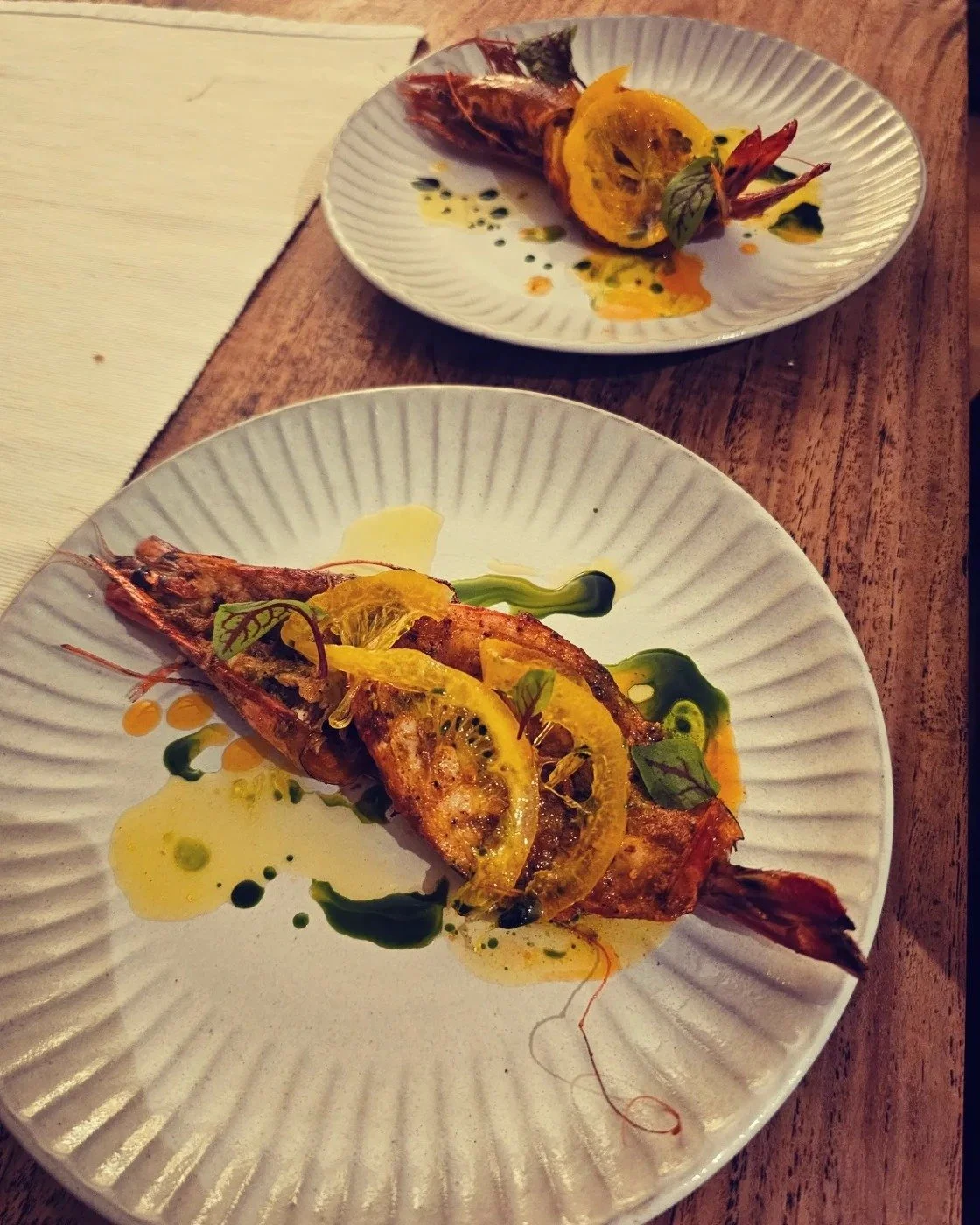 Two plates of grilled shrimp garnished with lemon slices, microgreens, and colorful sauces on a wooden table.