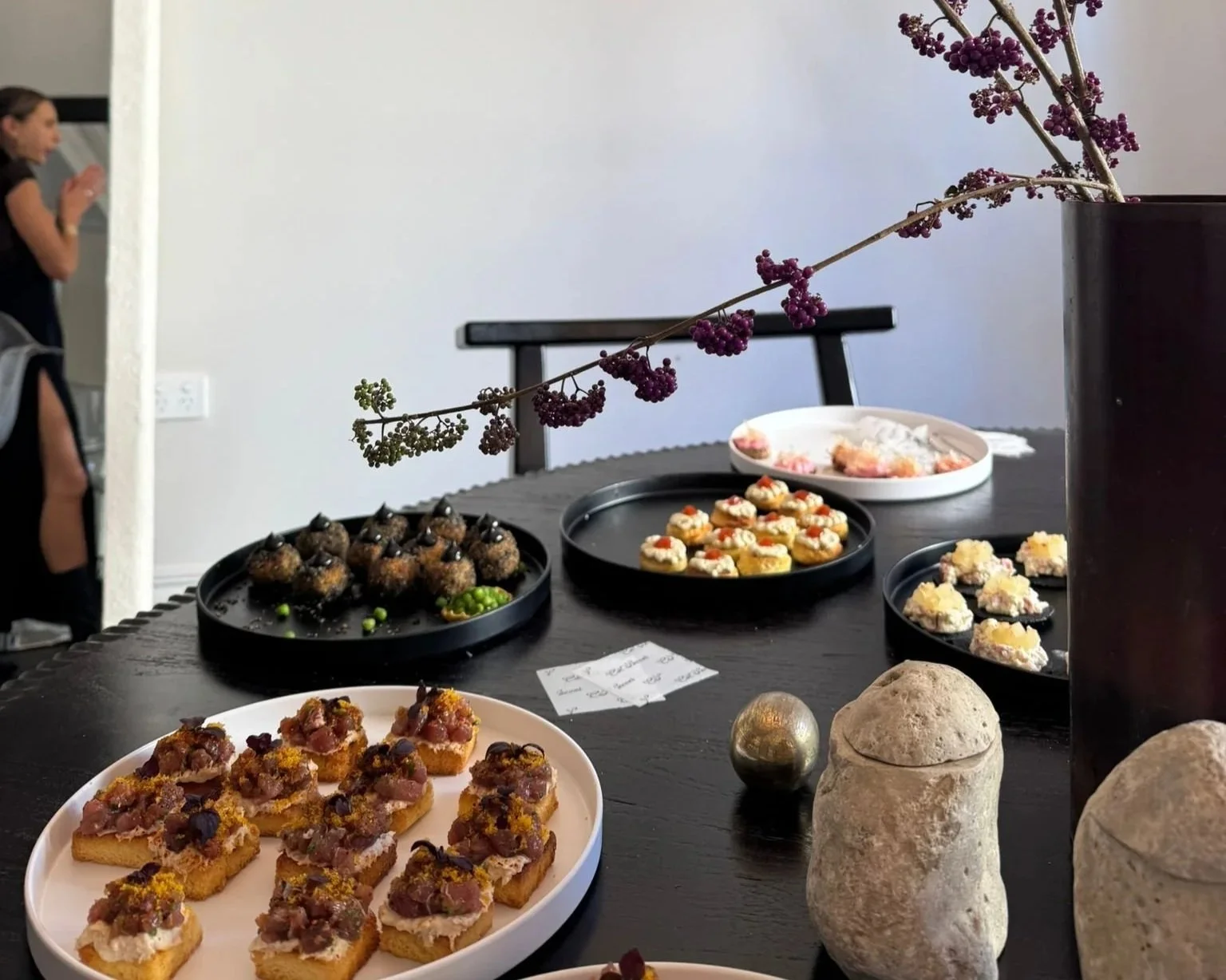 A table with various desserts and a centerpiece with a branch of purple berries in a black vase, with a person blurred in the background.