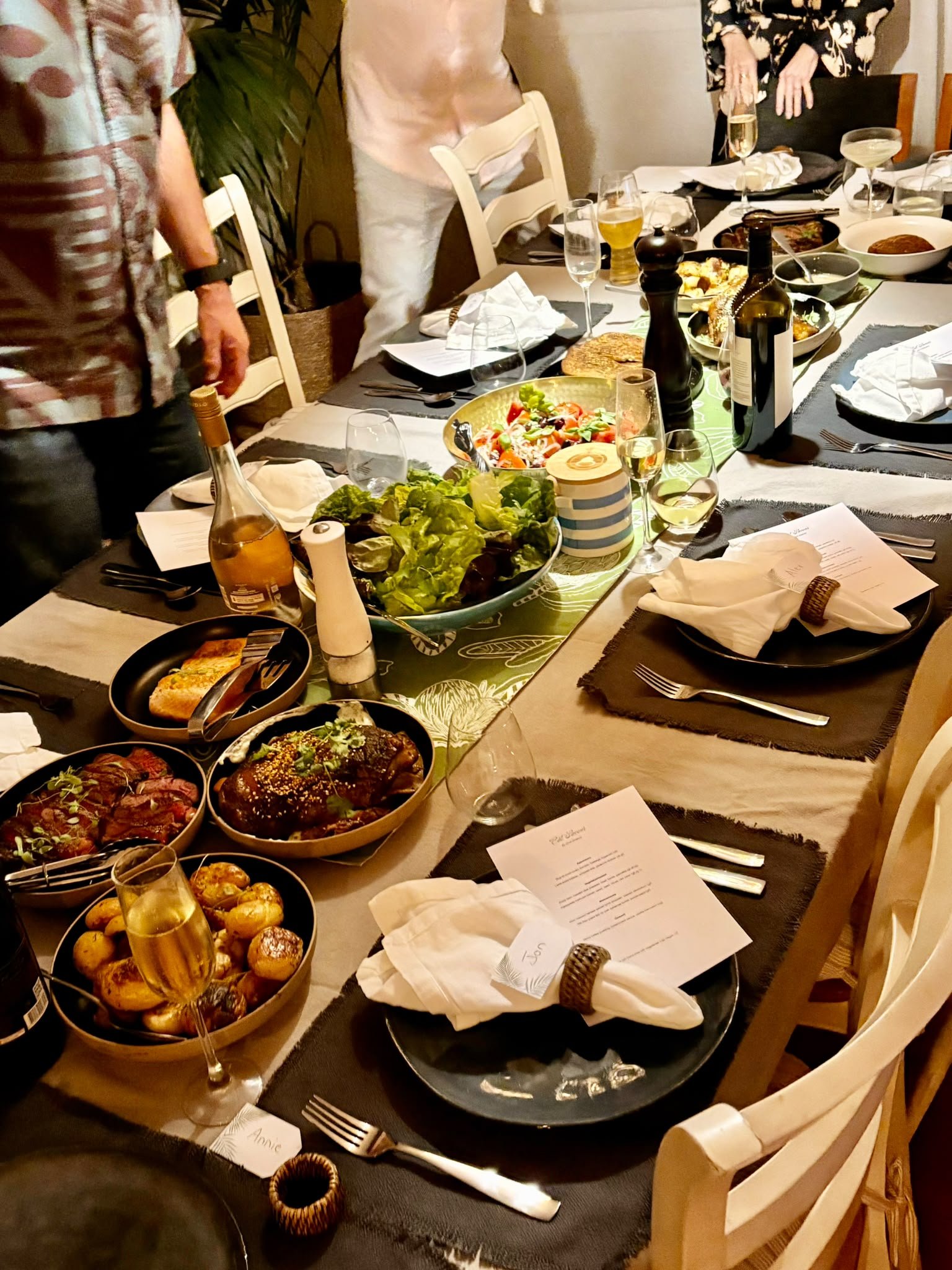 A dinner table set for a meal with various dishes, glasses of wine and champagne, and place settings with utensils and napkins. There are some people standing around the table.
