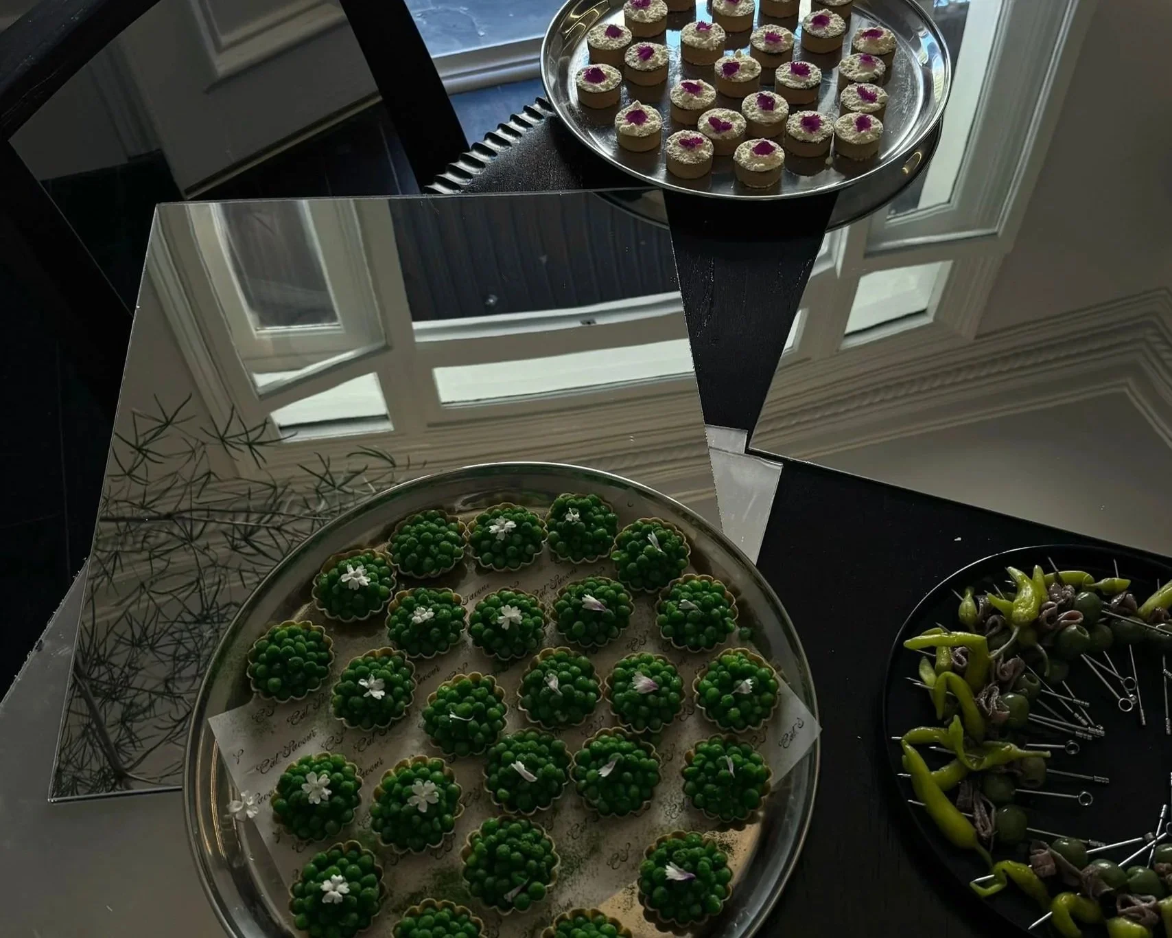A tray of small, round, green desserts decorated with white flowers and pink accents, placed on a silver platter. Above, there is a round metal tray with bite-sized desserts topped with white frosting and a purple flower. To the right, there is a black plate with various green and brown finger foods. The setting is near windows, with reflections of window frames in the background.