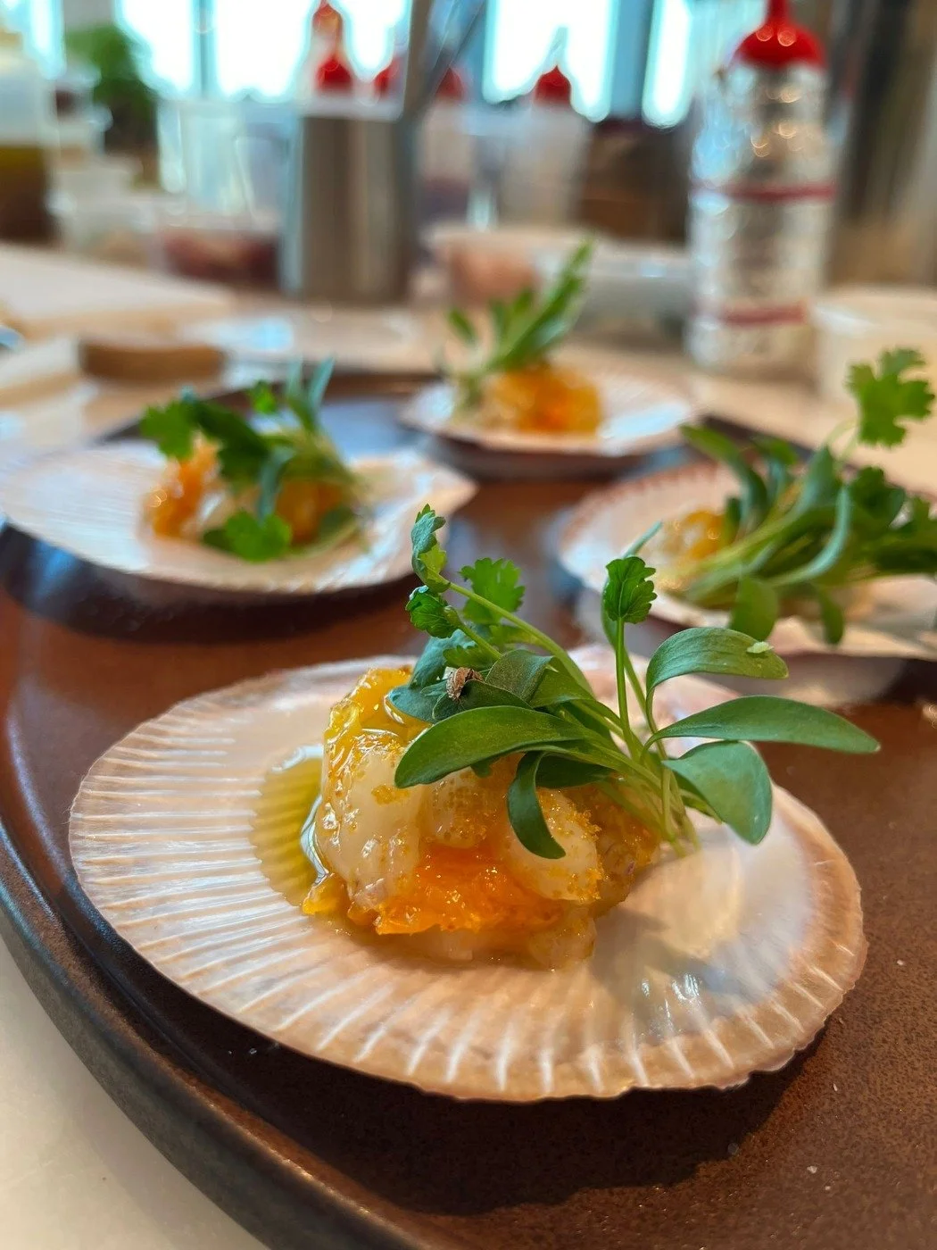 Close-up of a seafood dish with fresh herbs on a shell-shaped plate, garnished with greens, served on a dark tray, at a restaurant with a bright background.