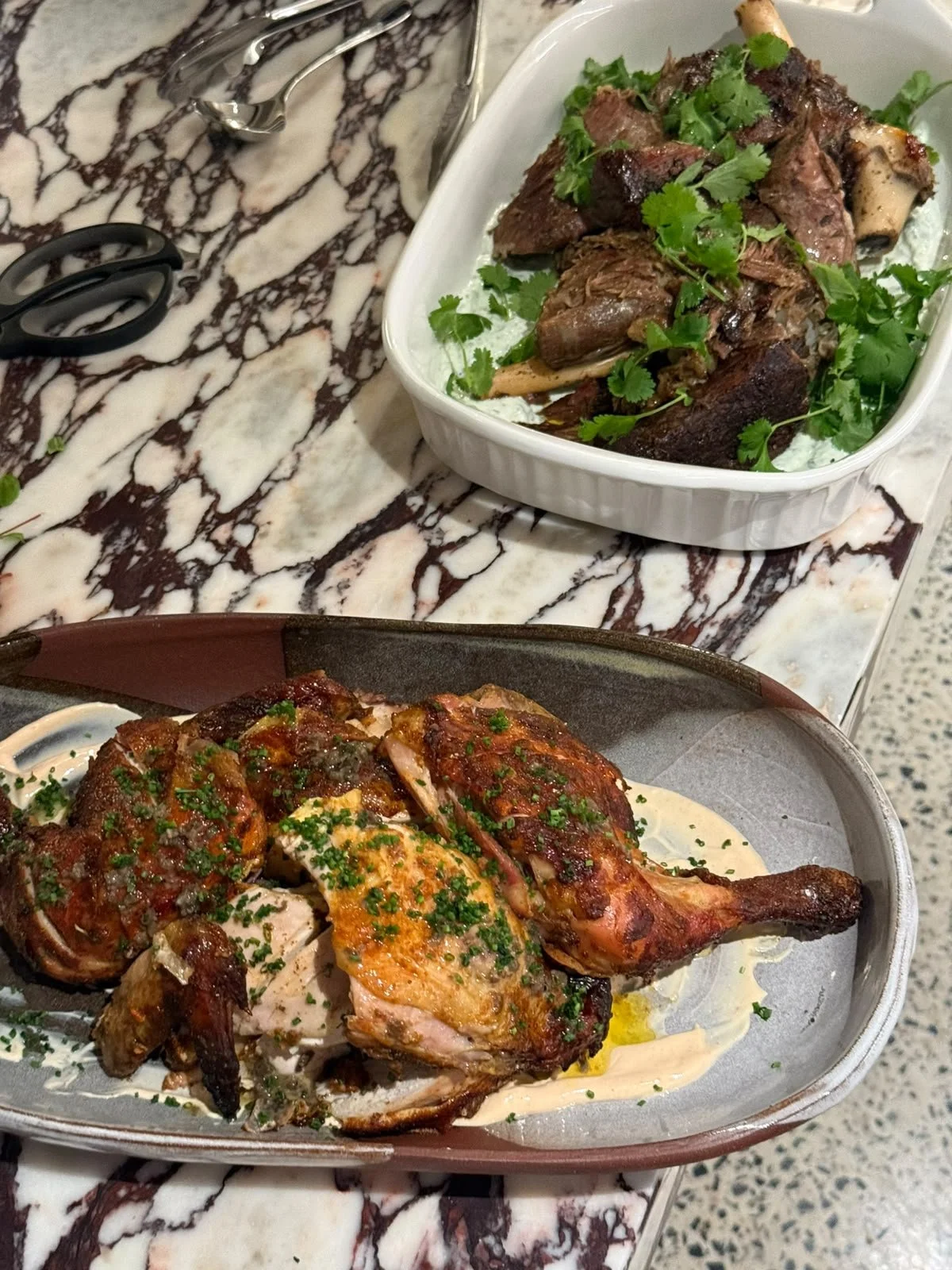 A roasted chicken half garnished with chopped herbs on a metal tray, and a dish containing cooked lamb shanks garnished with fresh herbs in a white oval dish, placed on a marble-patterned surface.