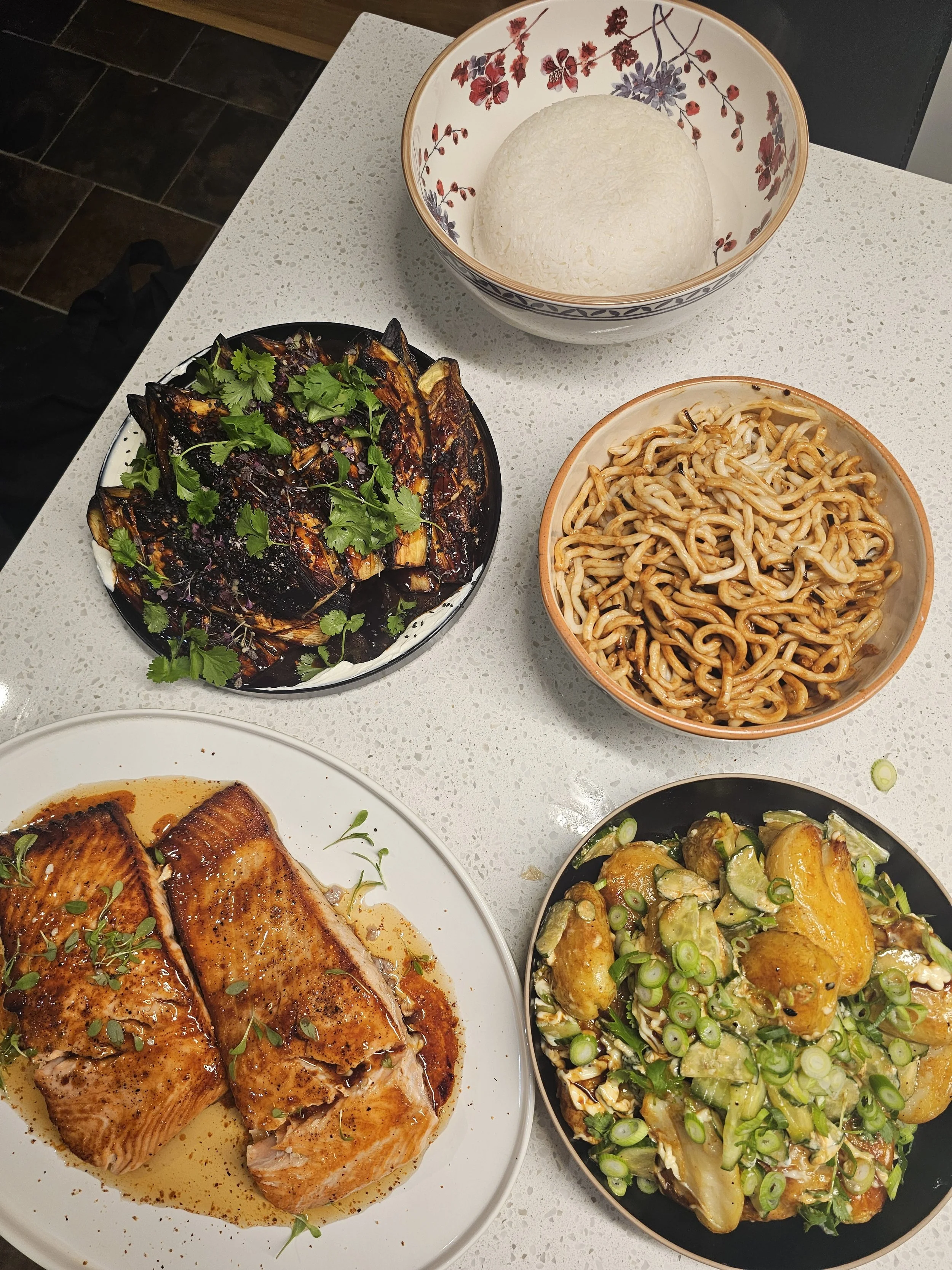 A dinner with several dishes on a white countertop, including a bowl of white rice, a plate of grilled salmon with herbs, a bowl of stir-fried noodles, and a dish of roasted vegetables topped with green onions.