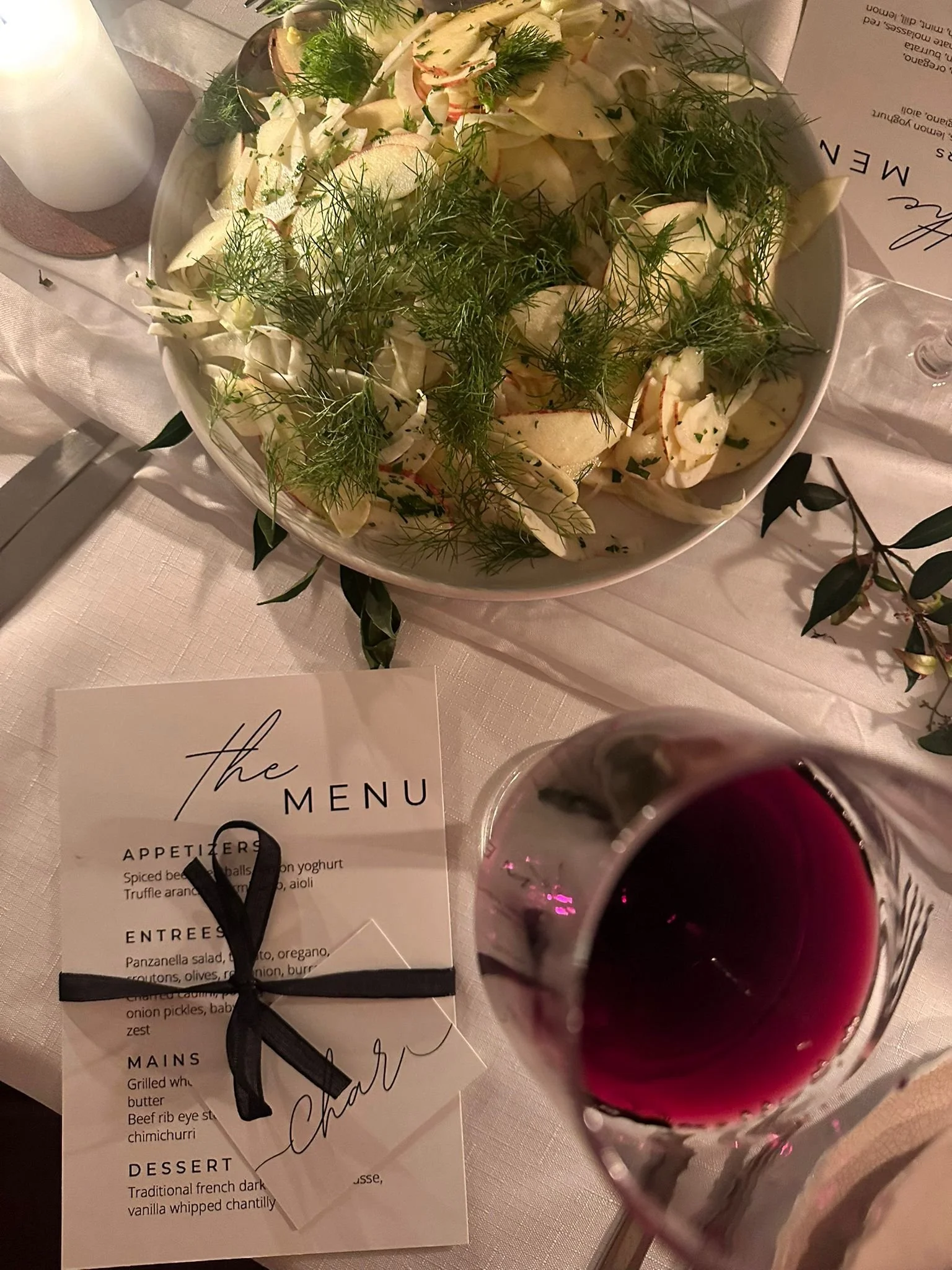 A white ceramic bowl containing a salad with mixed greens, fennel, and herbs. A glass of red wine is in the foreground. There is a menu card, a white vase, and some decorative greenery on the table.