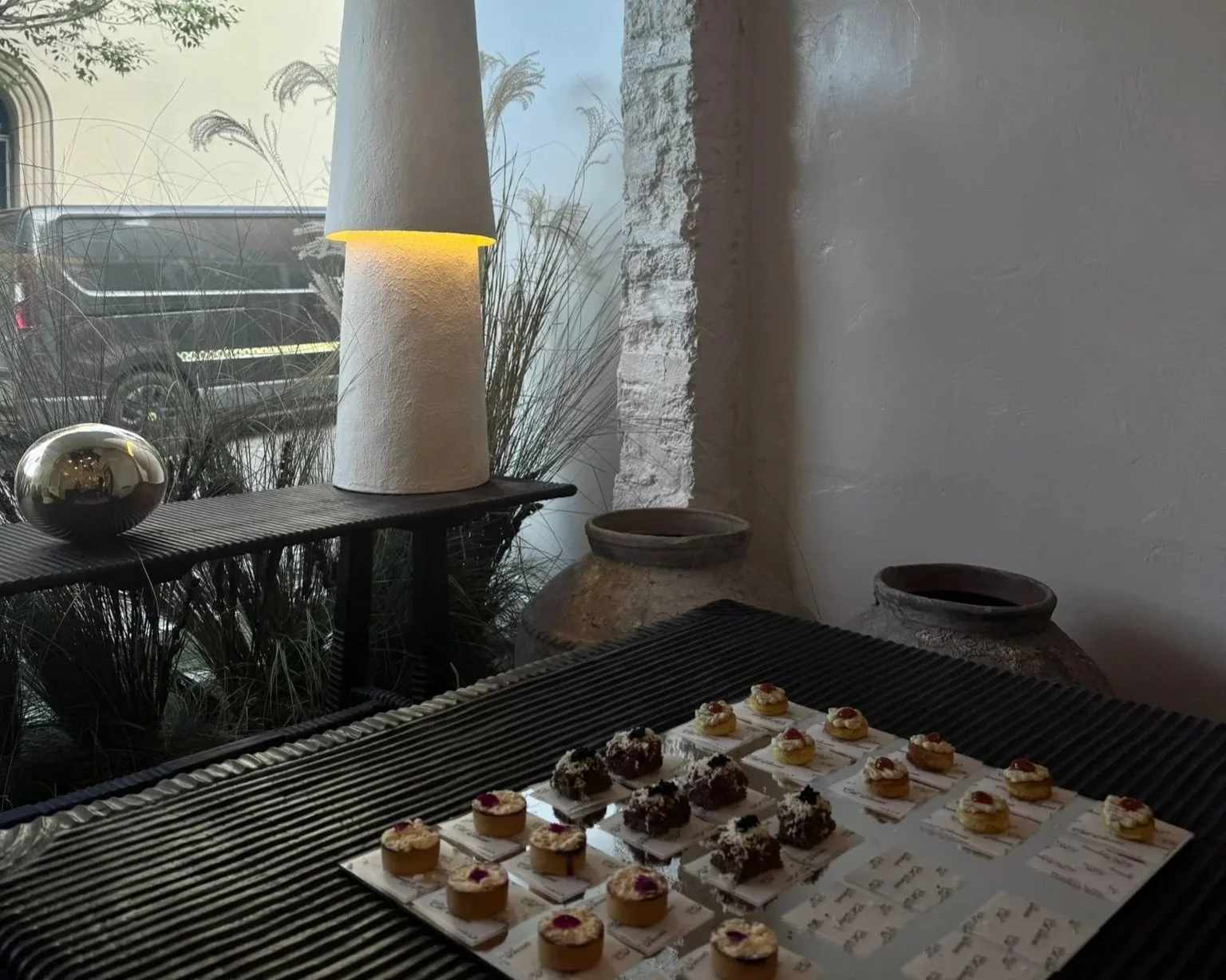 Display of assorted small desserts arranged on a table inside a modern, minimalist space with natural lighting, decorative pottery, and a lamp near a window.