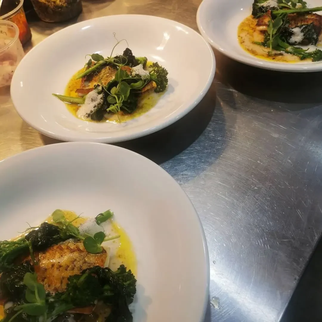 Three white bowls with plated fish and greens, on a shiny metal countertop.