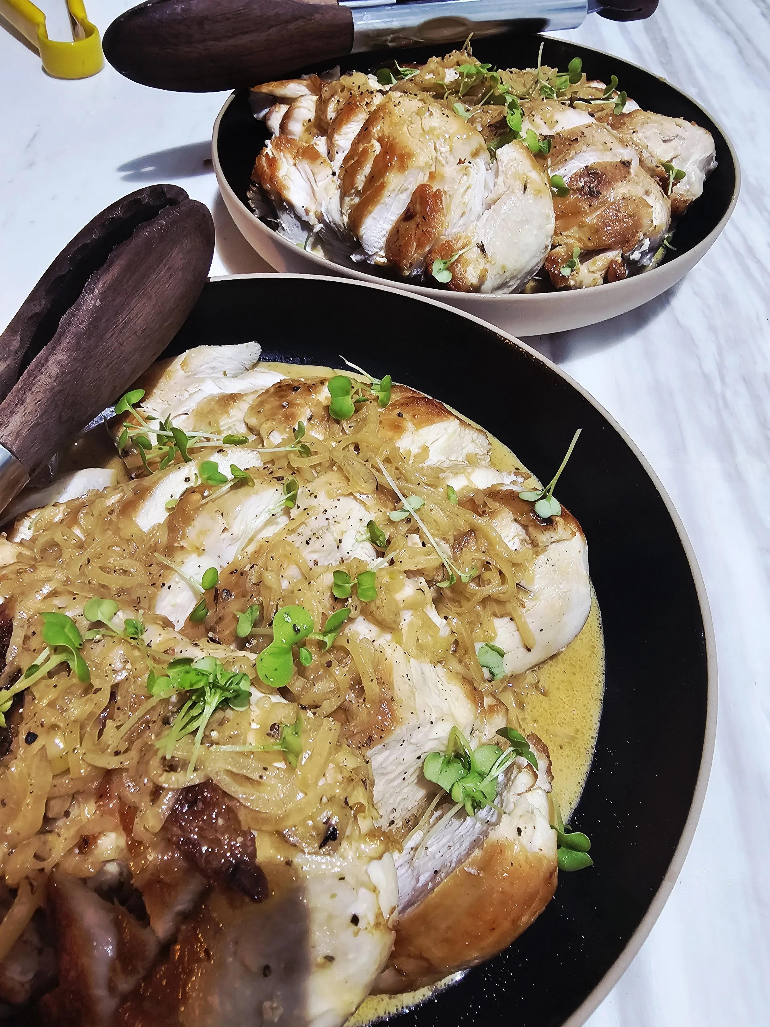 Baked whole chicken topped with caramelized onions and microgreens, served on a black plate.