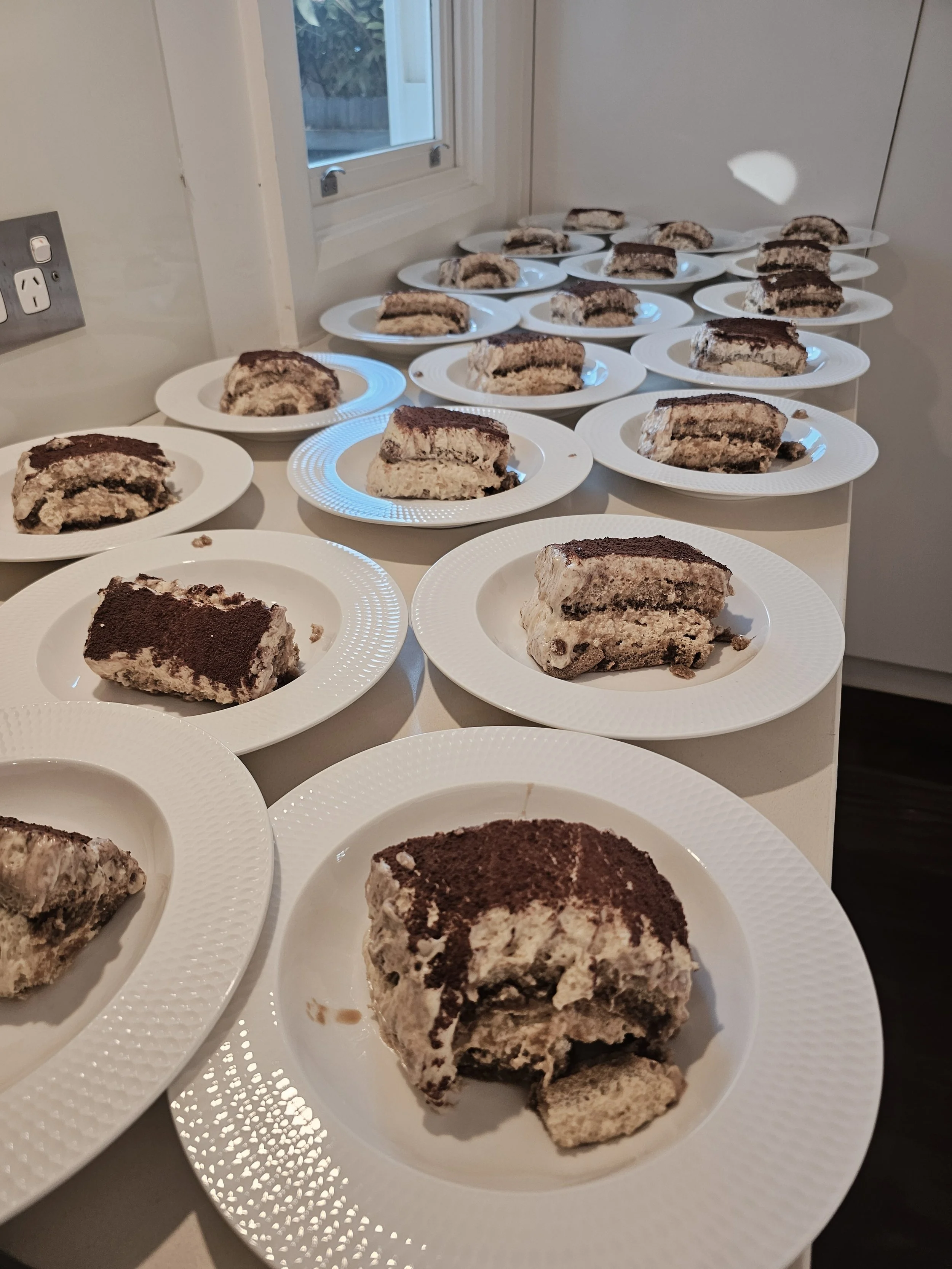 Multiple plates with slices of tiramisu dessert on a table, near a window in a kitchen.