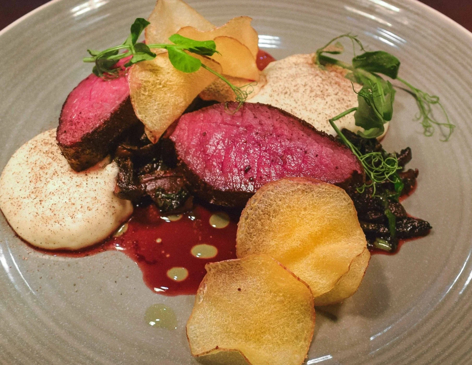 A plated dish featuring sliced medium-rare beef tenderloin, crispy potato chips, a dollop of smooth white puree garnished with sprinkled spices, microgreens, and a drizzle of sauce.
