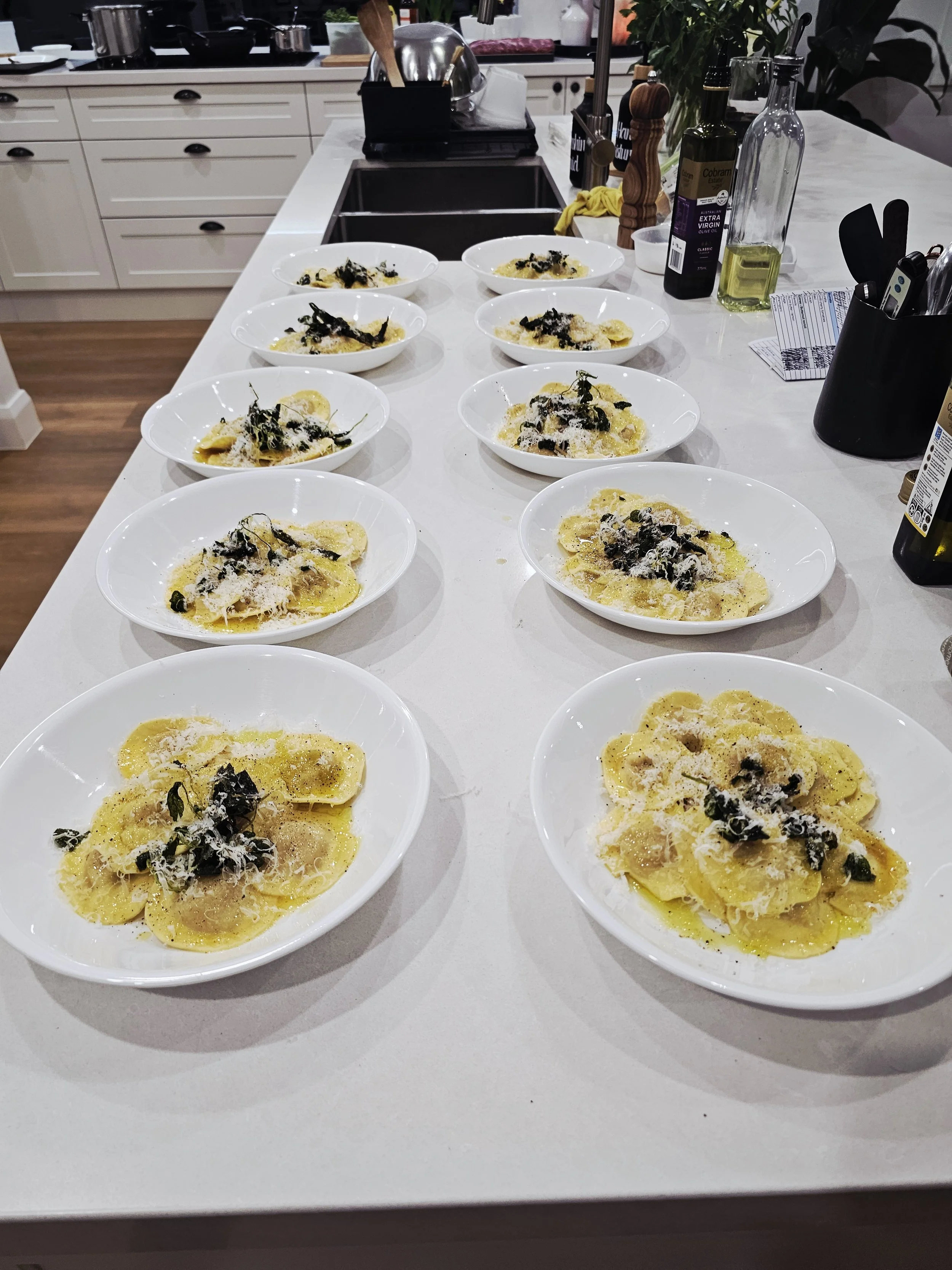 Several white bowls on a kitchen counter, each containing ravioli pasta garnished with black olives, grated cheese, and herbs. The kitchen background shows utensils, bottles, and a sink.