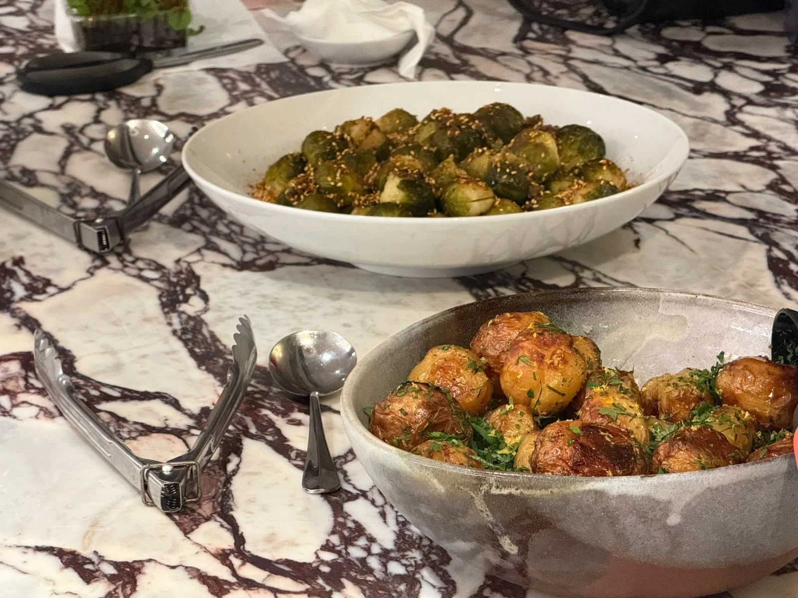 Two bowls of roasted Brussels sprouts and roasted potatoes on a marble table, with serving utensils, napkins, and a small dish of herbs or seasoning.