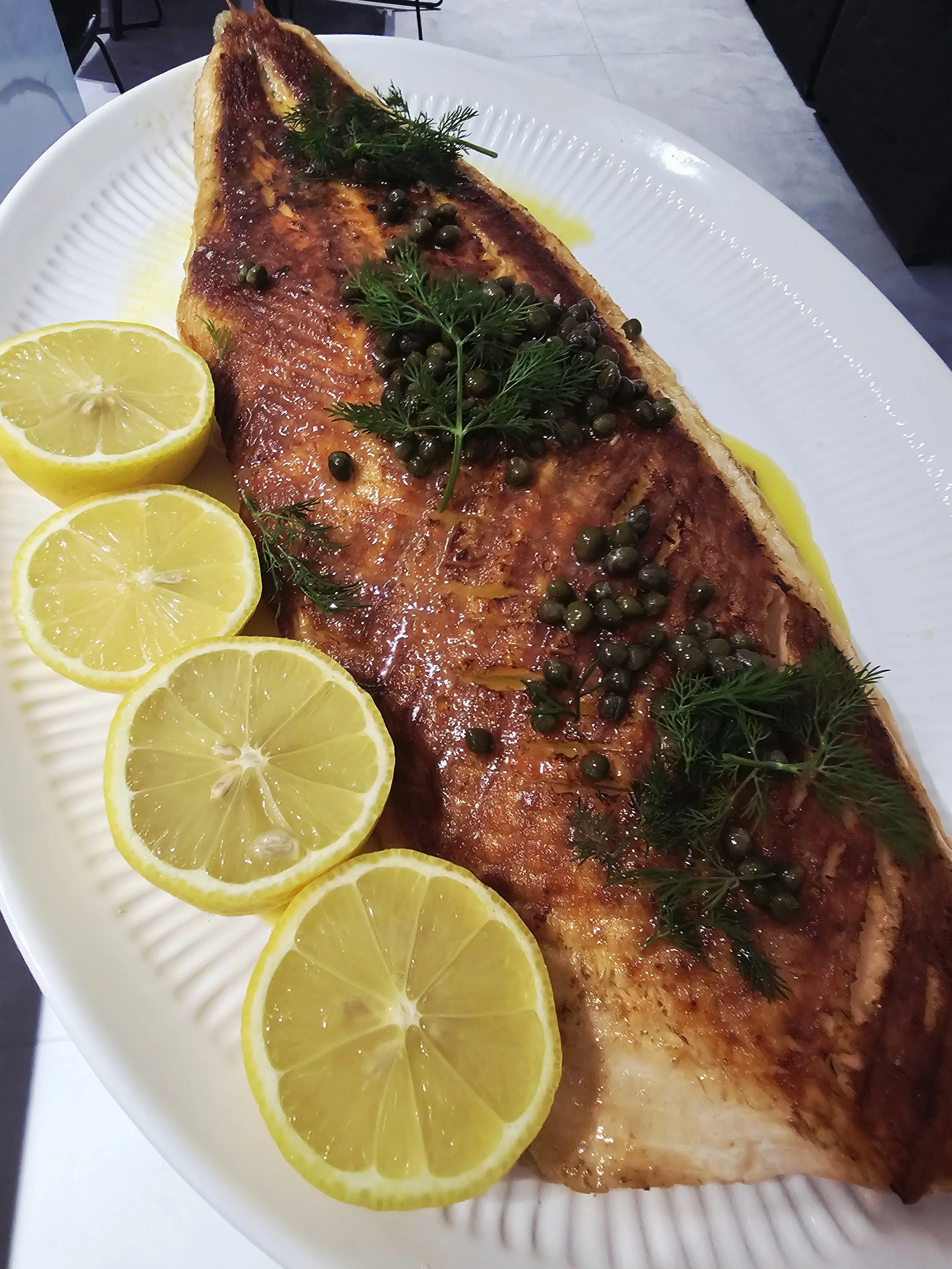 A cooked fish fillet garnished with black peppercorns and fresh dill, served on a white plate with sliced lemons.