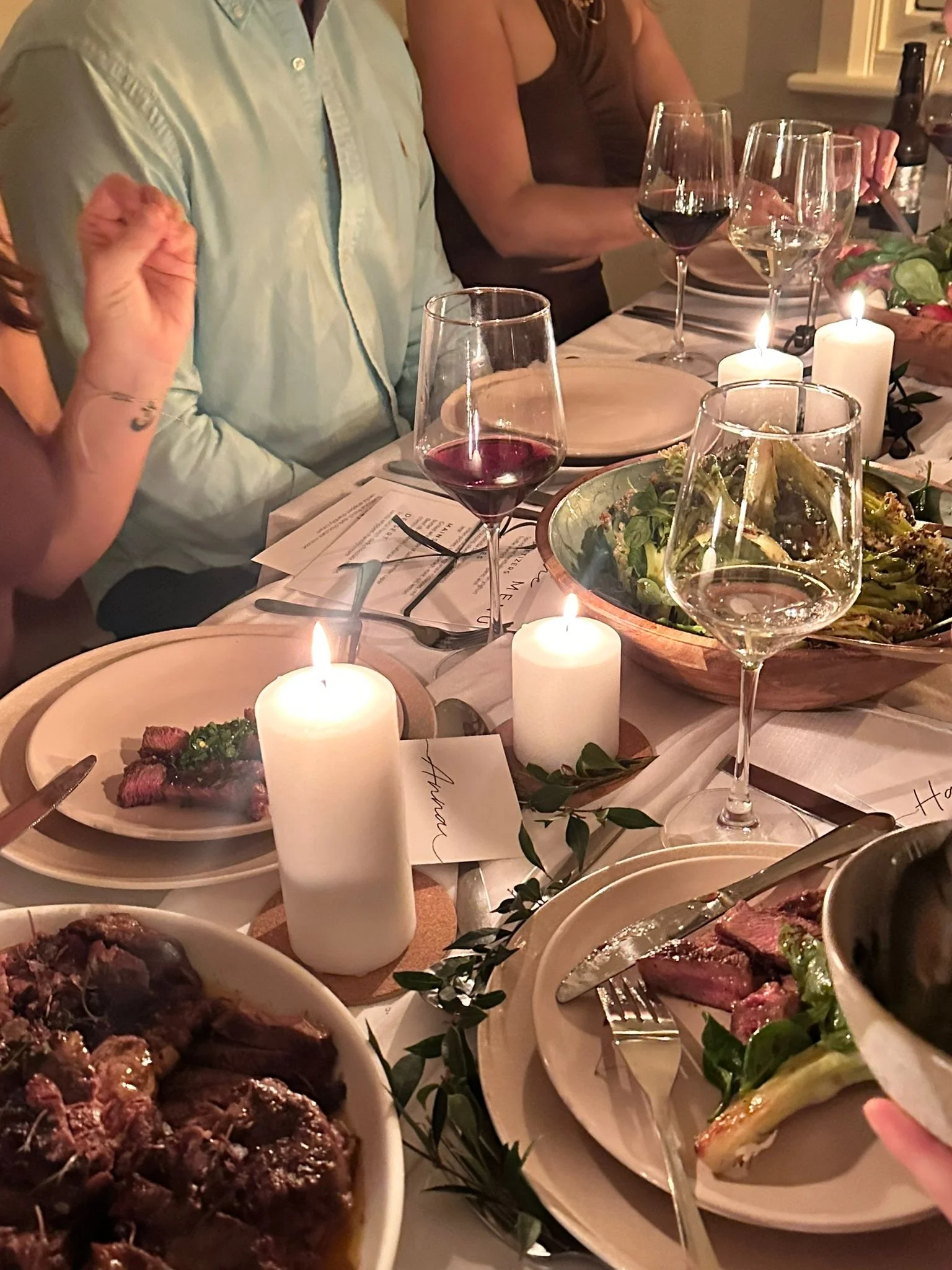 A dinner table set with candles, wine glasses, plates, and bowls of steak, salad, and other dishes during a family or social gathering.