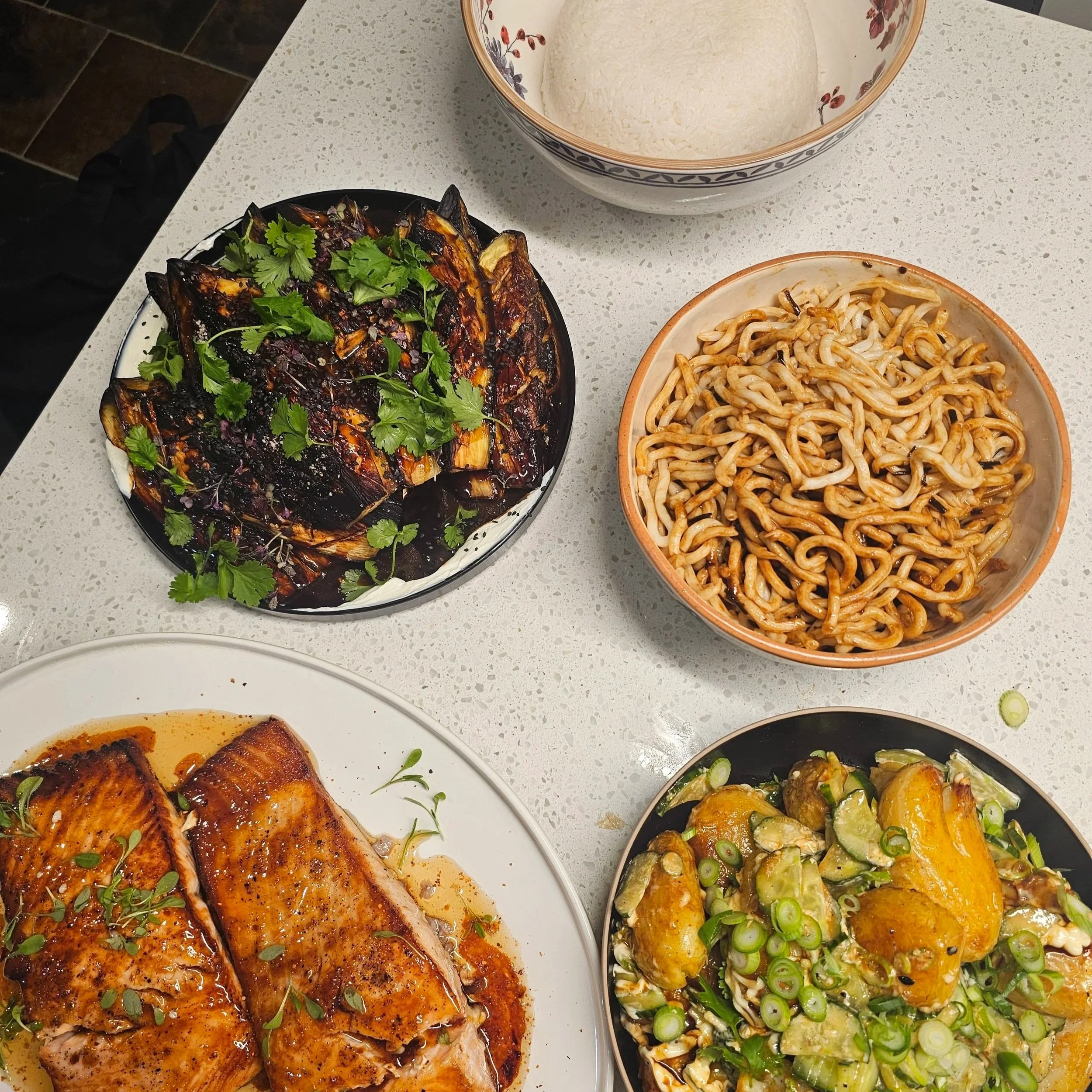 A table with various dishes, including grilled salmon topped with herbs, a bowl of noodles with sauce, and a bowl of roasted vegetables garnished with green onions and herbs.