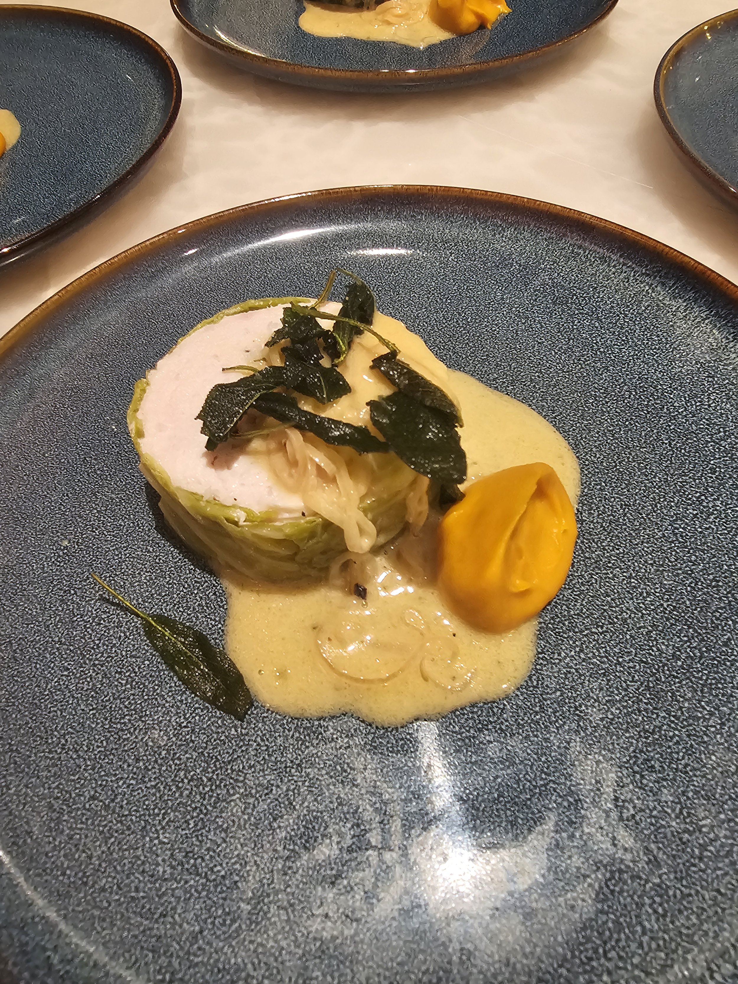 A culinary dish featuring a rolled piece of meat with a creamy yellow sauce, topped with dark herbs or seaweed, and accompanied by a dollop of bright orange puree on a textured dark blue plate.