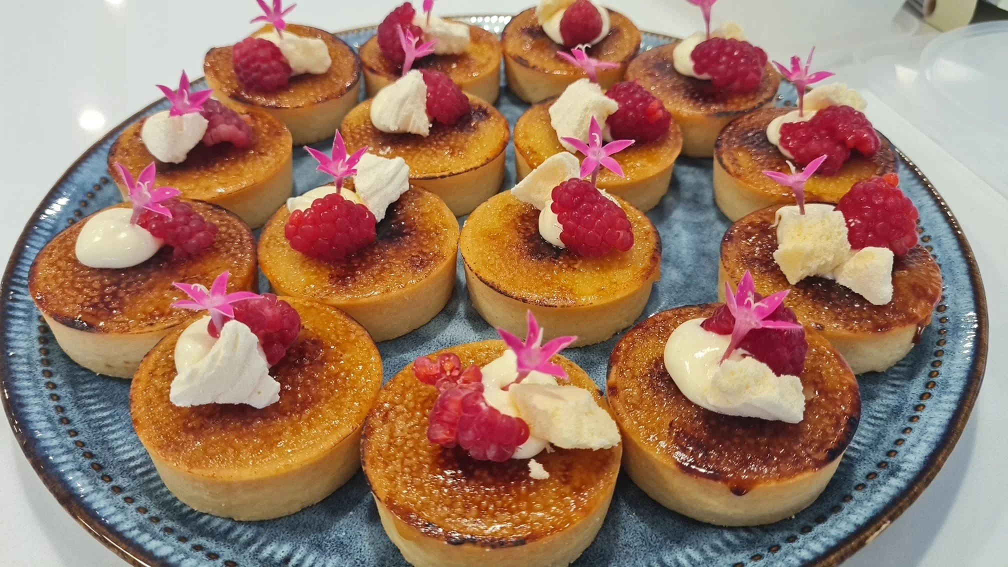 A plate of small, round crème brûlée desserts topped with raspberries, white cream, and pink edible flowers