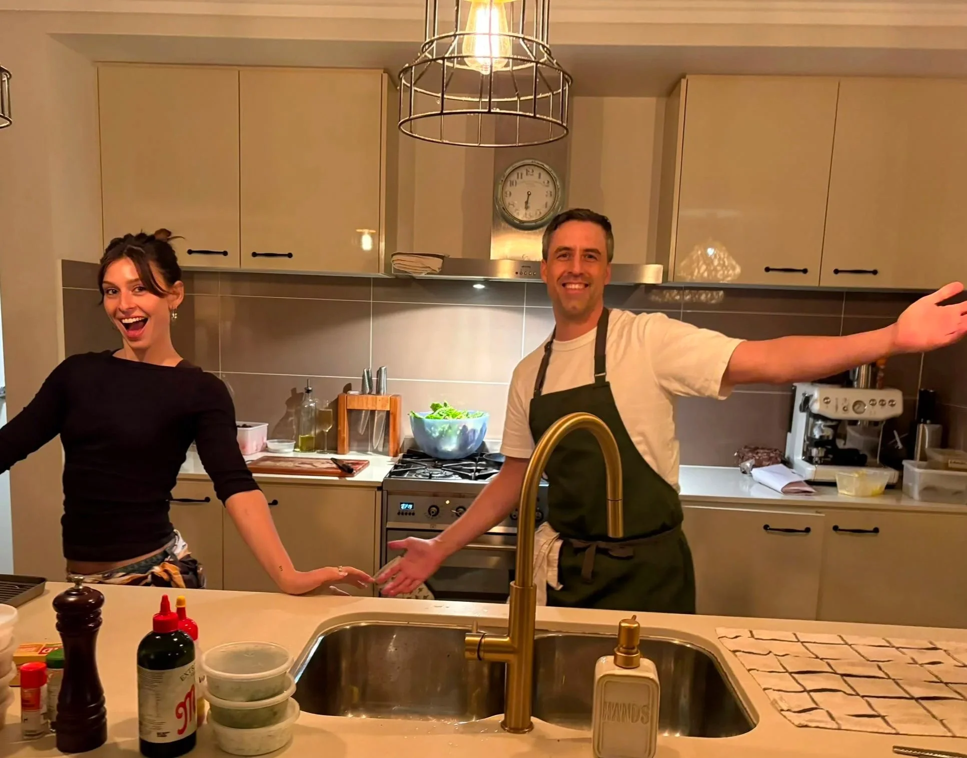 A man and a woman in a kitchen, smiling and holding hands, with various kitchen utensils and ingredients on the counter.