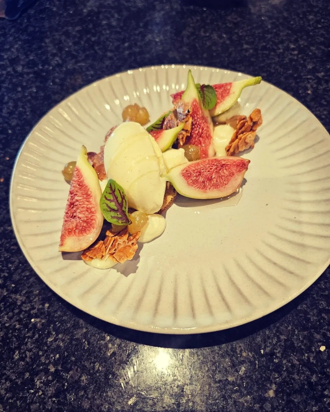 A dessert plate with fresh figs, walnuts, cream, and microgreens on a white plate with a textured rim, placed on a black granite countertop.