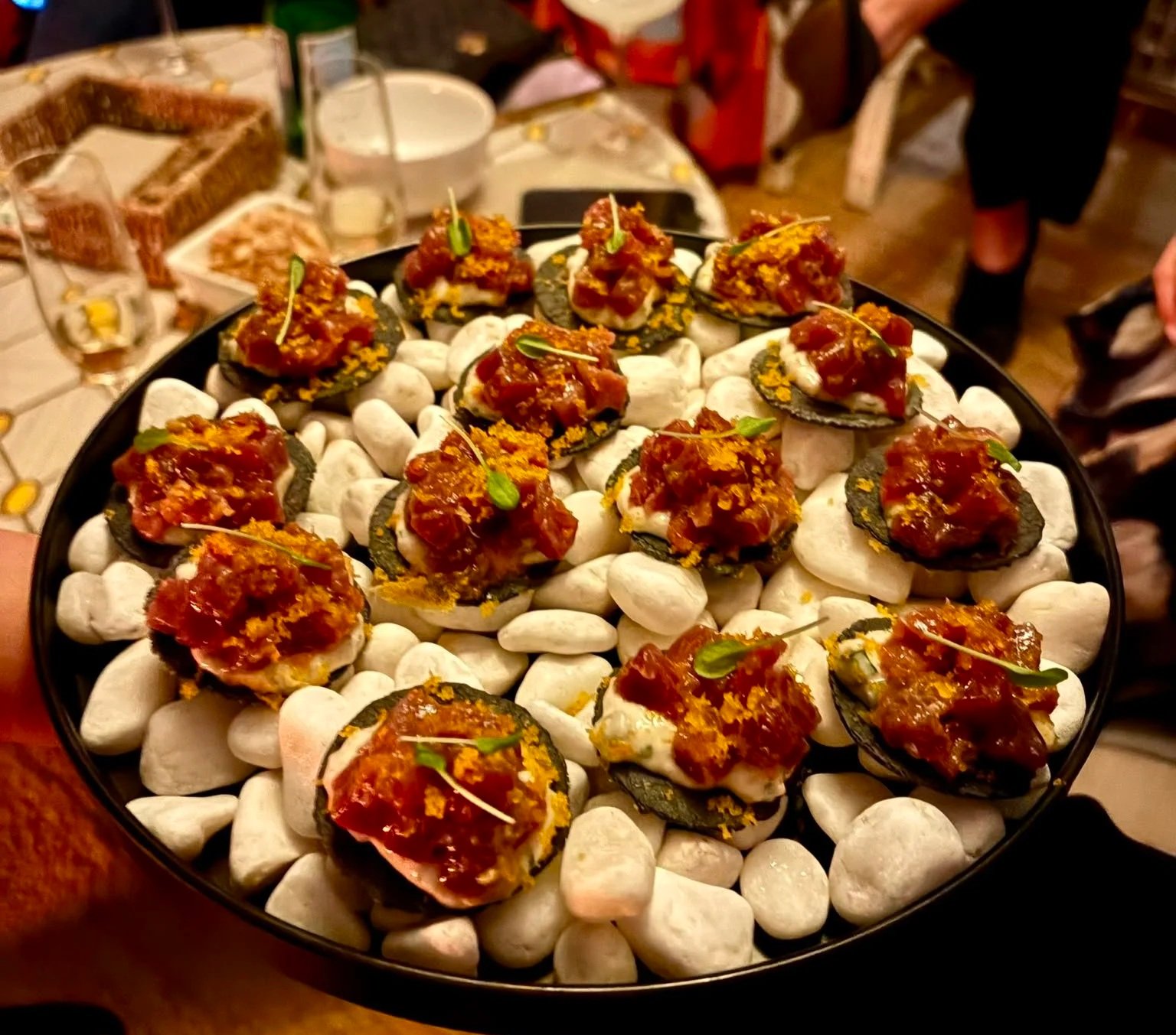 A platter of stuffed mushrooms topped with sauce and garnished with microgreens, arranged on white stones at a dining table with drinks and utensils in the background.