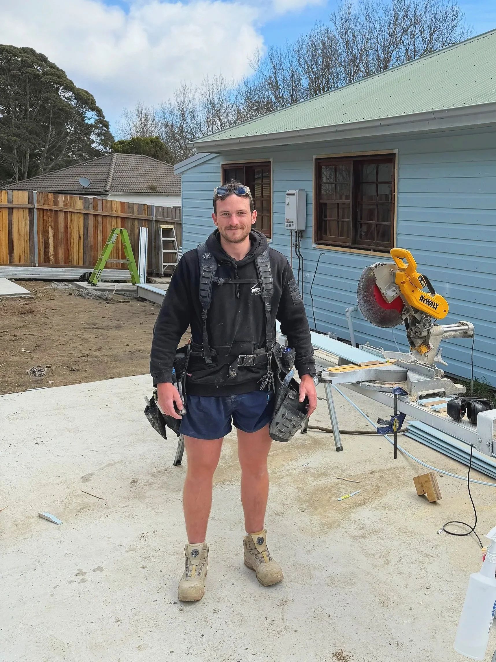 A man in construction gear standing on a concrete foundation in front of a blue house during daytime. He is holding a tool belt, wearing beige work boots, shorts, and a black jacket, with safety glasses on his head. Construction materials and tools are visible around him, including a miter saw, a ladder, and a wooden fence in the background.