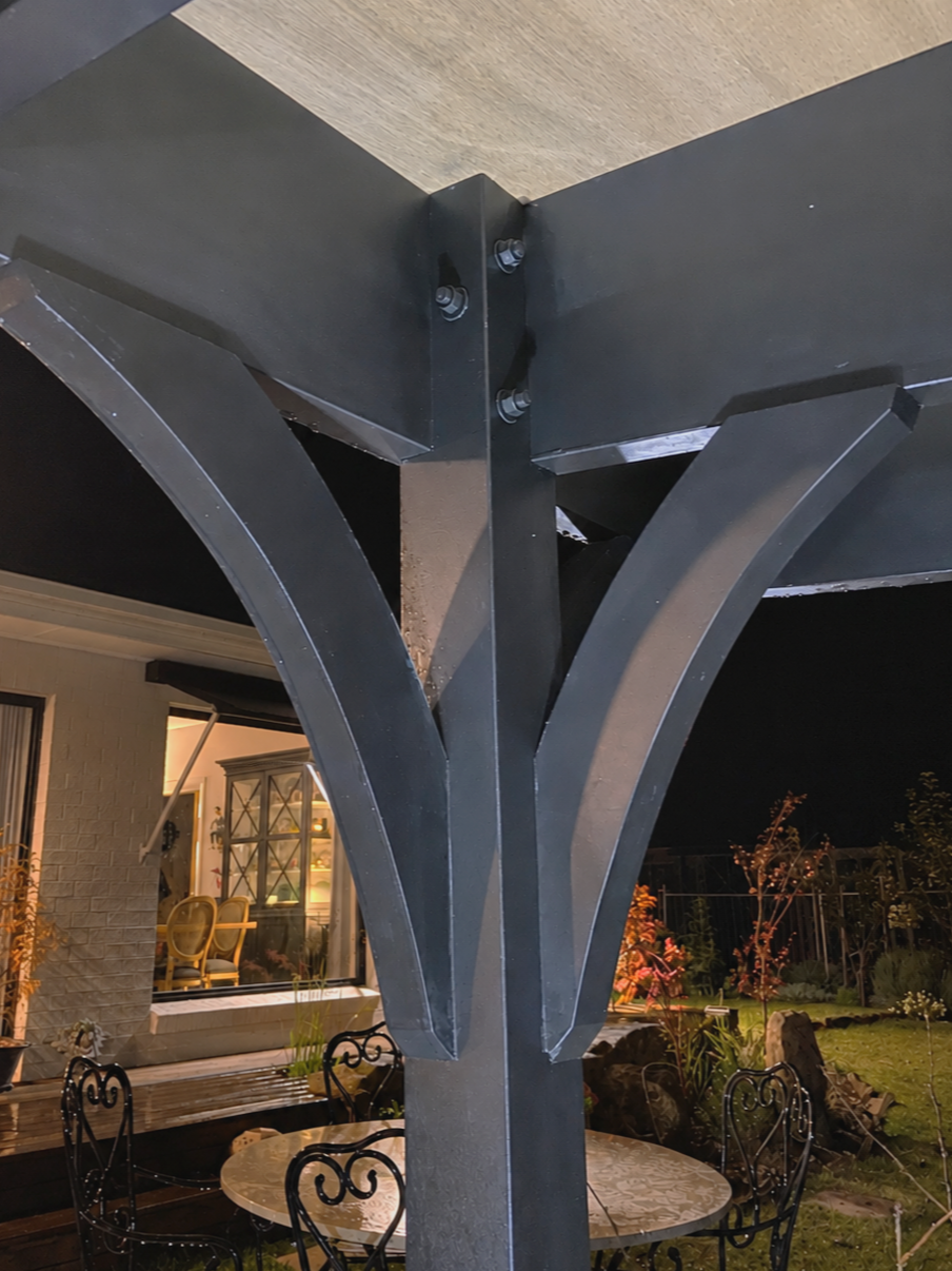 Close-up of a black outdoor patio table with four ornate black chairs around it, situated near a house with a window and a garden at night.